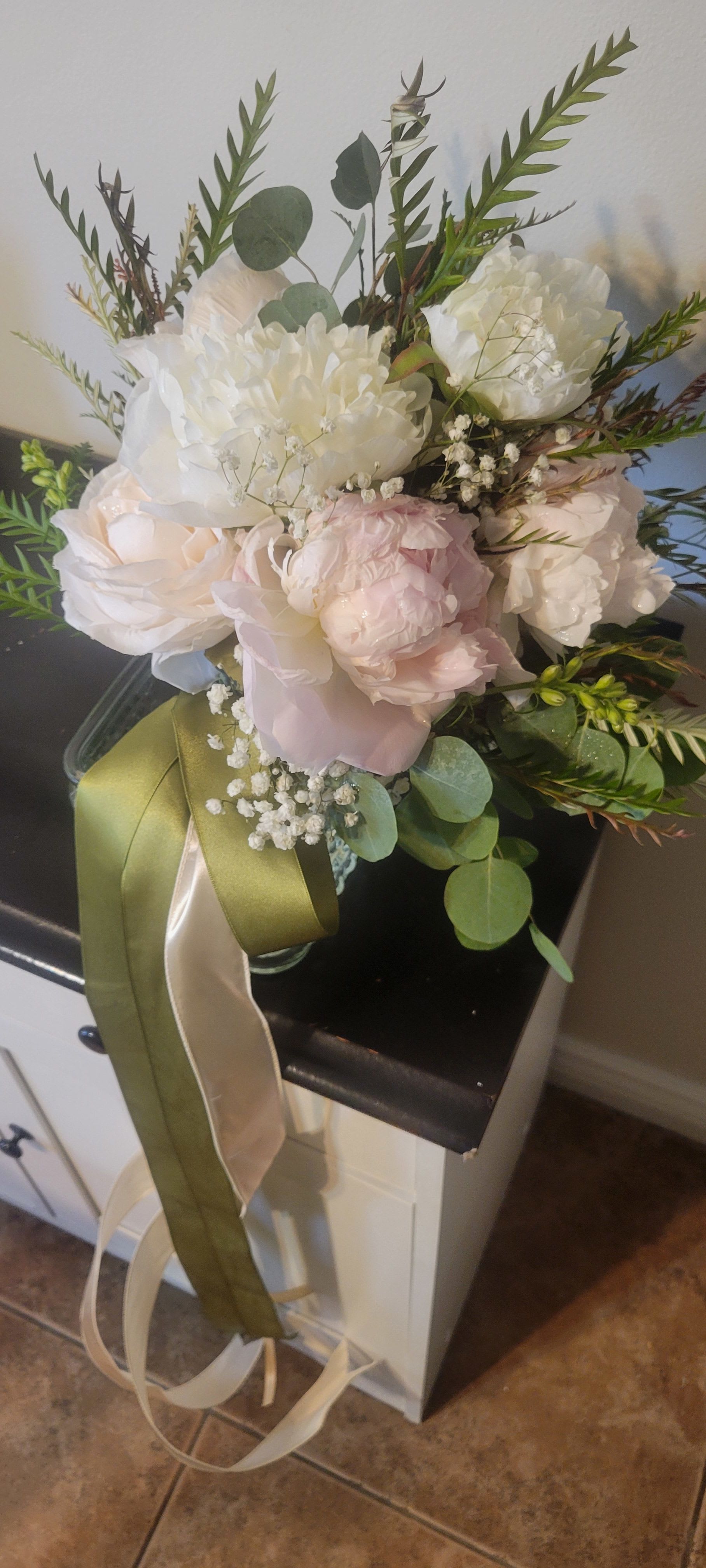 Bridal Peonie Bouquet by Dixie's Flowers - This is a soft and romantic bridal bouquet. It features cream and blush peonies, baby's breath, and greenery like eucalyptus and ferns. The bouquet is tied with long olive green and ivory ribbons that hang over the side, giving it an elegant and natural look.  