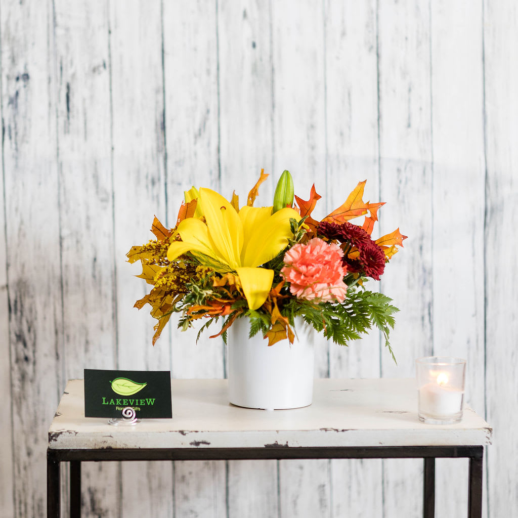 Flaxen - Vivid and bright ~ a fall arrangement including an asiatic lily, carnations and preserved fall leaves, all arranged in a white ceramic container.  Approximate Dimensions: 9&quot; H x 9&quot; W