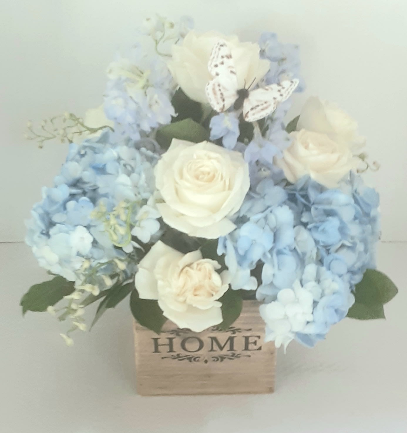 Where the Heart Is - Arranged beautifully in a modern all wood cube,with white roses, blue delphinium and blue hydrangeas and accented with lemon leaf,is perfect for many occasions, including a welcome to your new home! 