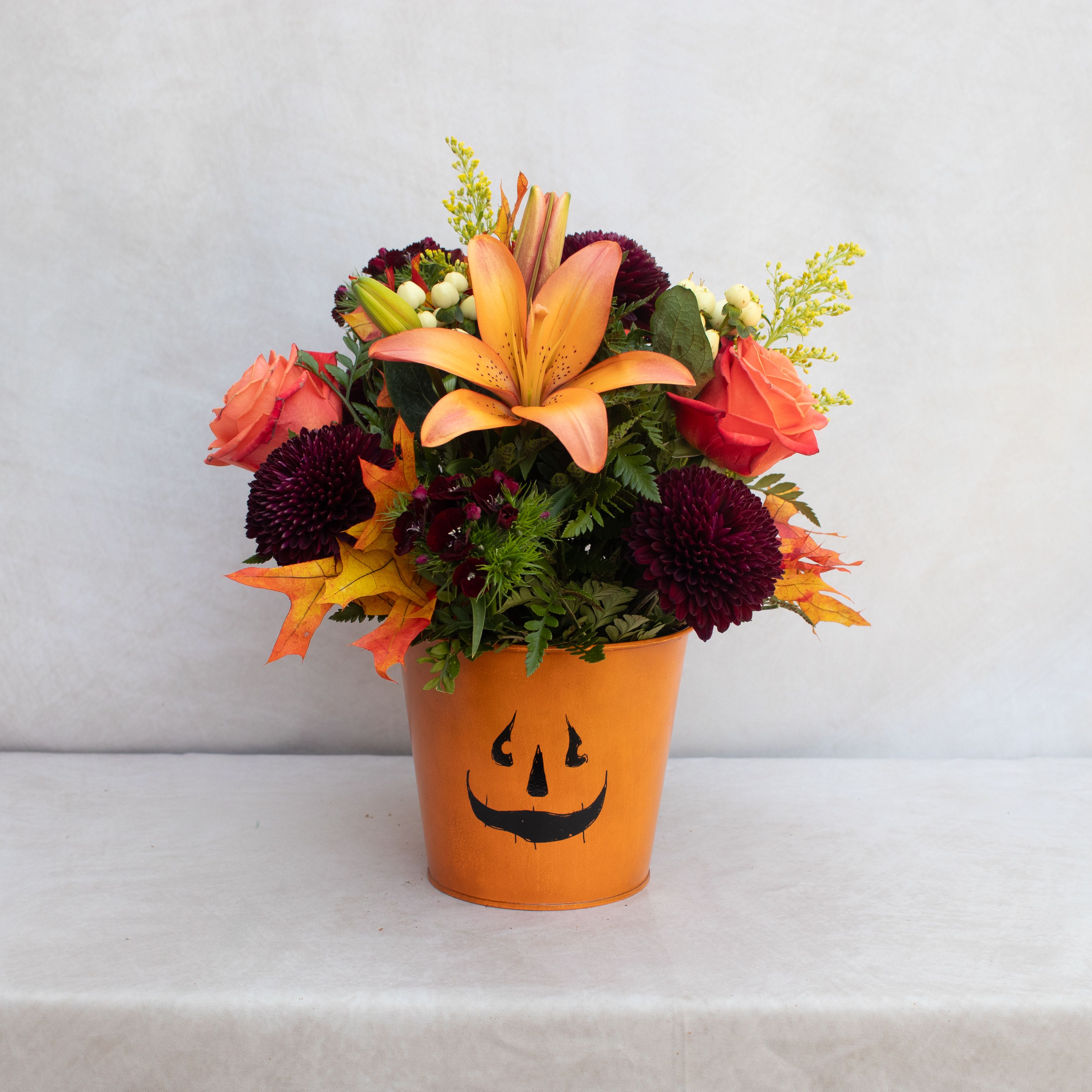 HALLOWEEN TIN  HALLOWEEN TIN  - A MIXURE OF FALL FLOWERS IN AN ORANGE METAL TIN WITH A SPOOKY GRIN!  TO INCLUDE SOME OF THESE FLOWERS: LILIES, ROSES, MUMS, LEAVES, SOLIDAGO, DIANTHUS, BERRIES, POMPS. ARRANGEMENT IS ABOUT 8X8 DESIGNED.