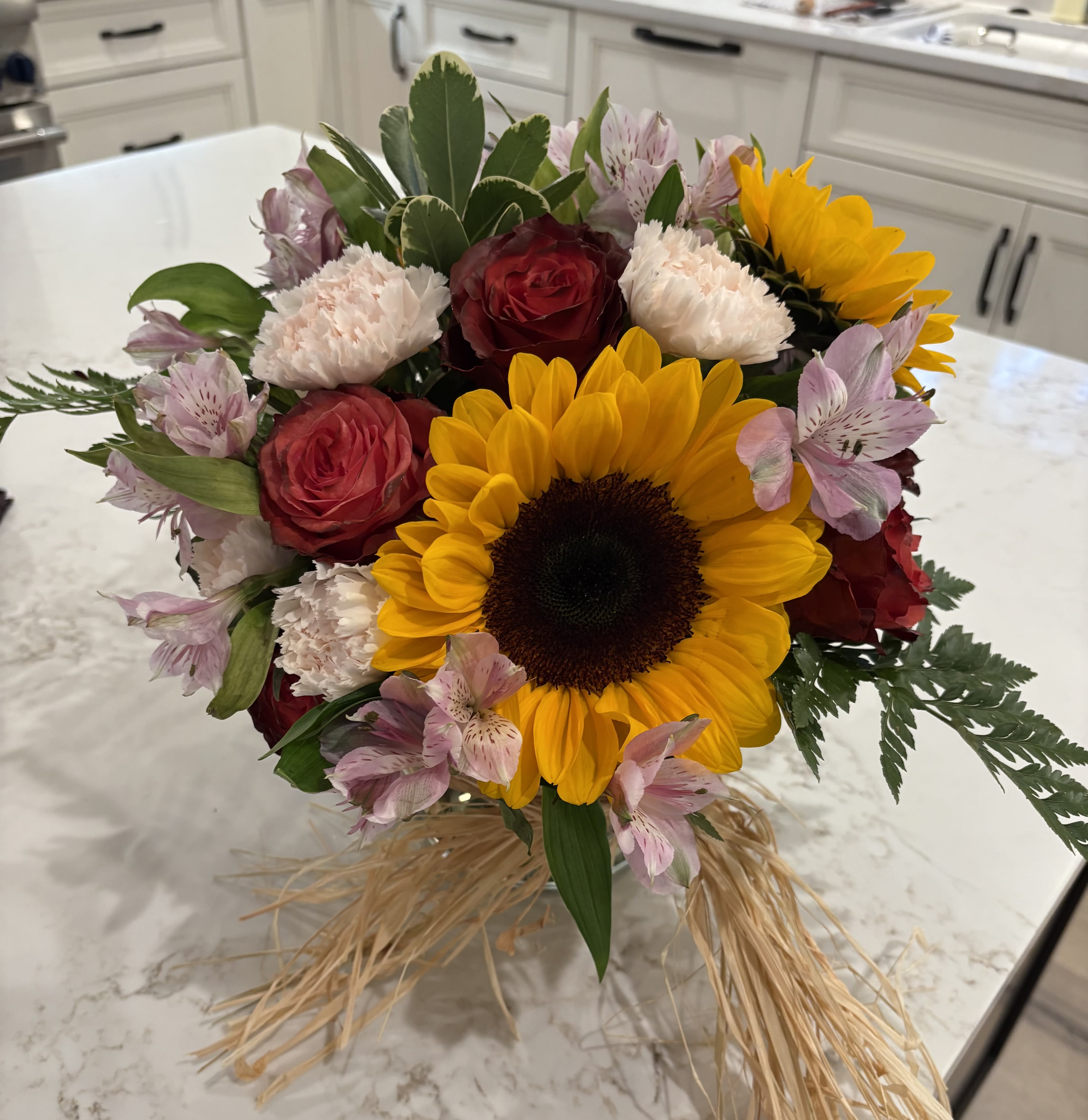 Hayride Bouquet - Brighten any day with this cheerful mix of blooms! The Hayride Bouquet features golden sunflowers, radiant red roses, soft pink alstroemeria, and delicate carnations — all tied together with natural raffia for a rustic, farm-fresh touch. Perfect for autumn celebrations, thank-you gifts, or just because, this bouquet brings warmth, joy, and a little country charm to every occasion.  Best For: Birthdays, fall gatherings, “thinking of you” moments, and kitchen table smiles.