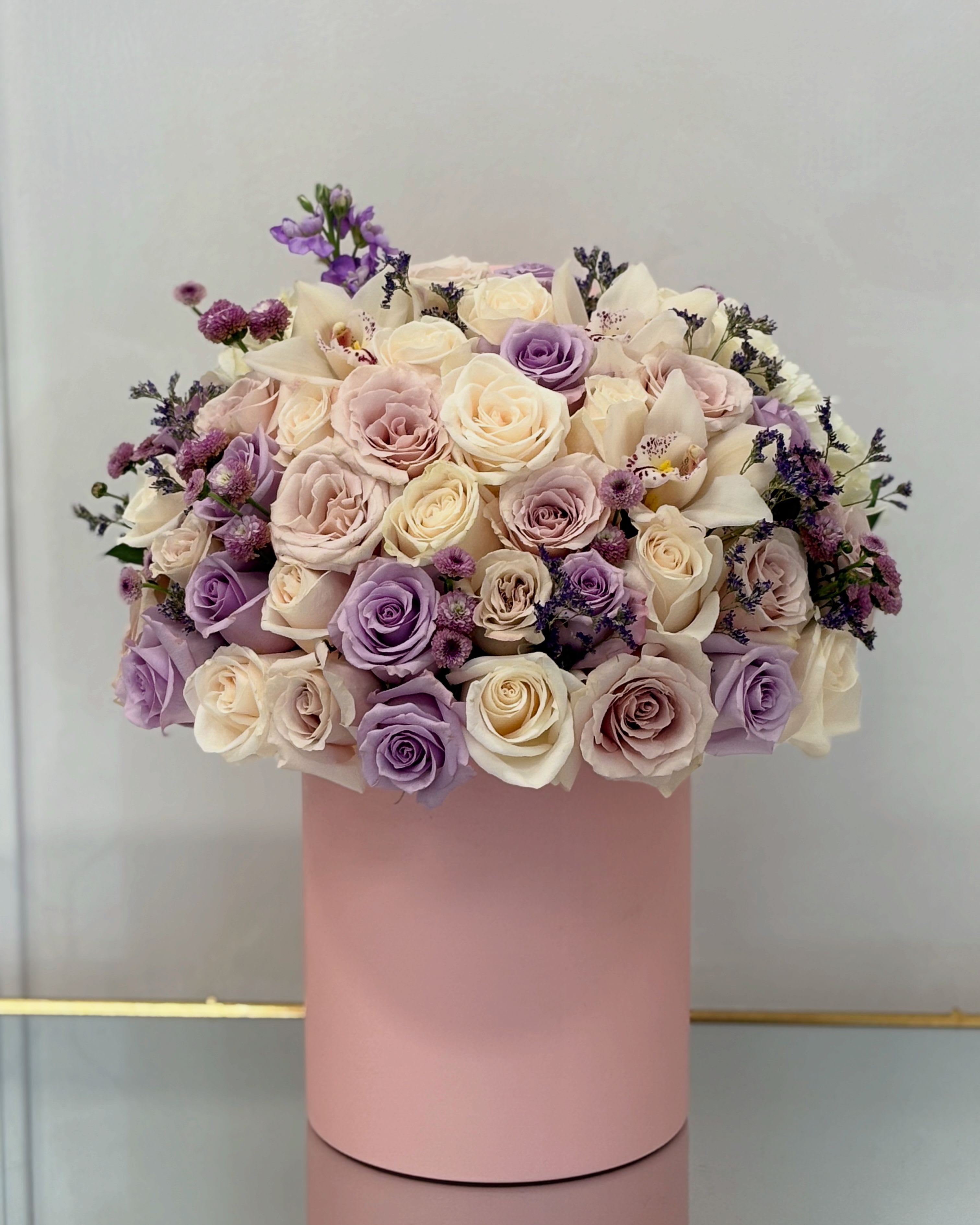 Lavender Whisper Hat Box By VLM Flowers - Soft, serene, and effortlessly elegant, Lavender Whisper is a dreamy floral arrangement featuring a blend of cream, blush, and lavender-hued roses. Accentuated with delicate sprigs of purple statice and whimsical asters, this arrangement is presented in a pale blush pink hatbox, evoking a sense of gentle romance and timeless charm. Perfect for expressing gratitude, celebrating delicate moments, or simply bringing peace and beauty into any space.