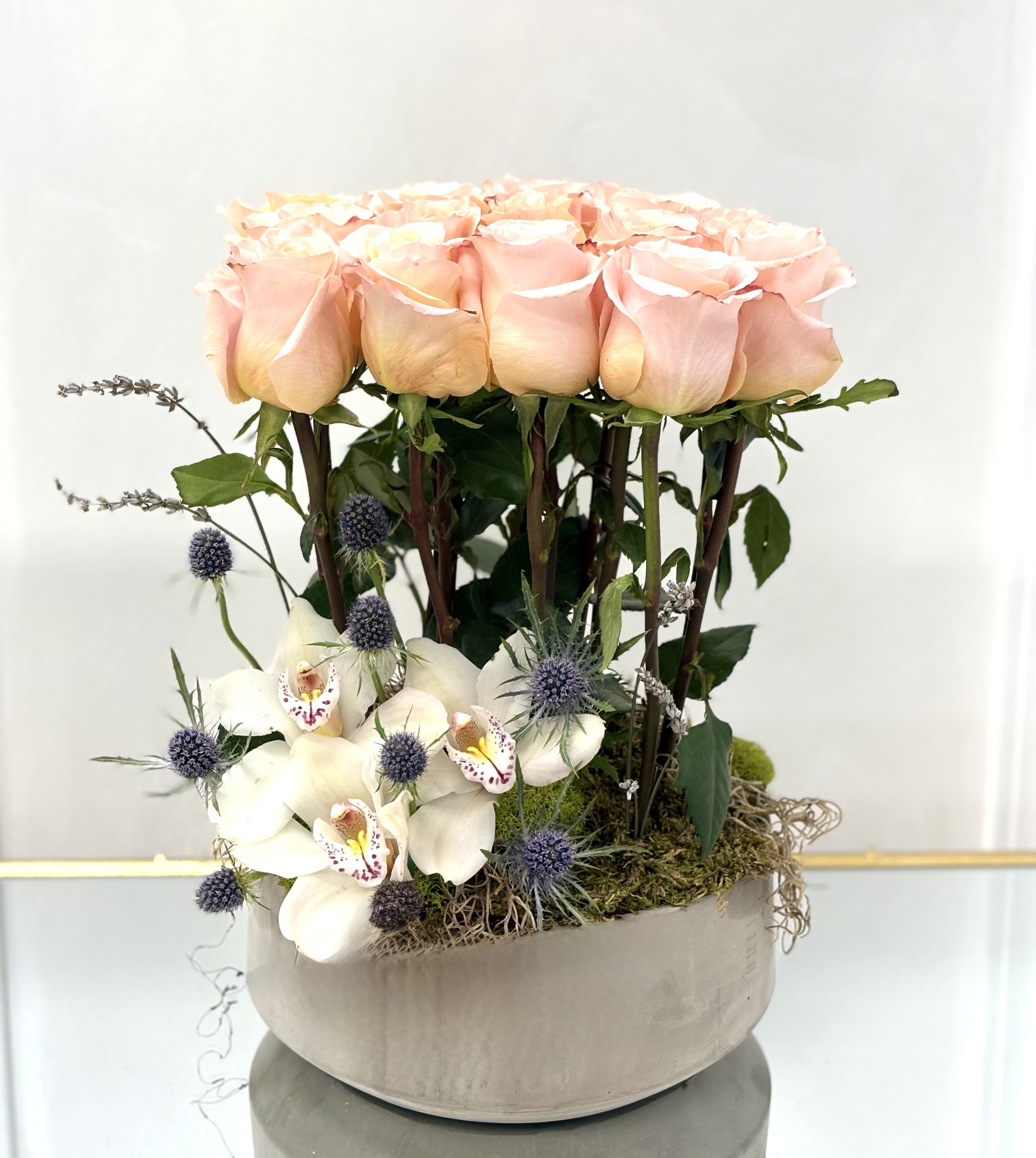 Rose Canopy By VLM Flowers - A modern garden dream, Rose Canopy features a tall cluster of soft peach roses rising like a floral forest above a bed of white cymbidium orchids, blue thistle, and natural moss. This structured and artistic arrangement evokes a woodland fantasy with refined elegance, perfect for high-style events, gallery displays, or as a memorable gift for someone who appreciates bold, sculptural beauty.