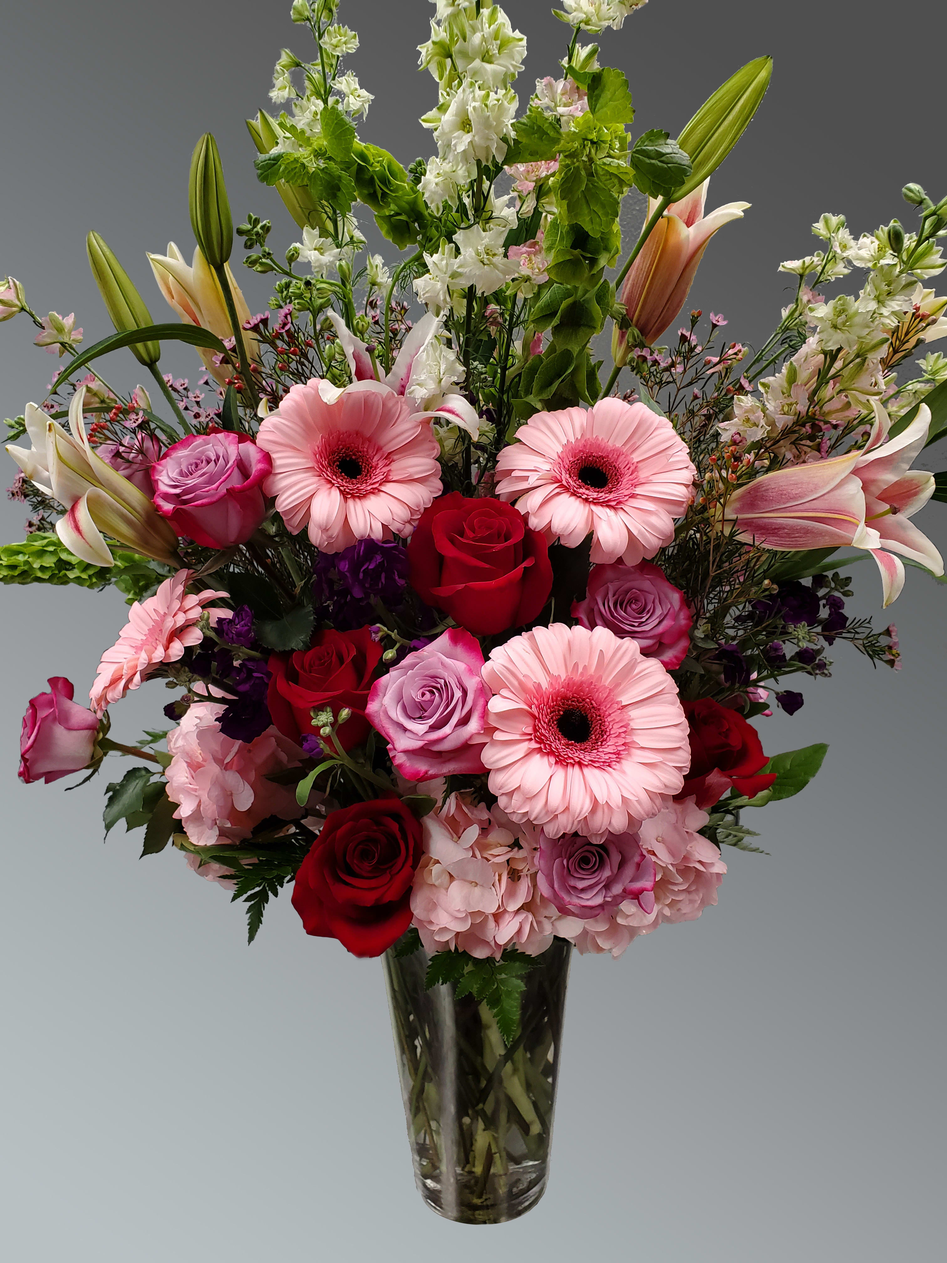 Aphrodite   - Aphrodite From our exclusive collection, Aphrodite is a breathtaking floral masterpiece designed to captivate. This stunning arrangement features an elegant blend of:  Lilies  Tulips  Larkspur  Gerberas  Lavender and red roses  White hydrangea  Purple lisianthus  Purple Monte Cassino asters  Stock  Lush mixed greenery  All artfully arranged in a clear glass vase, this design is exceptional in every way — a true expression of beauty, grace, and timeless elegance.