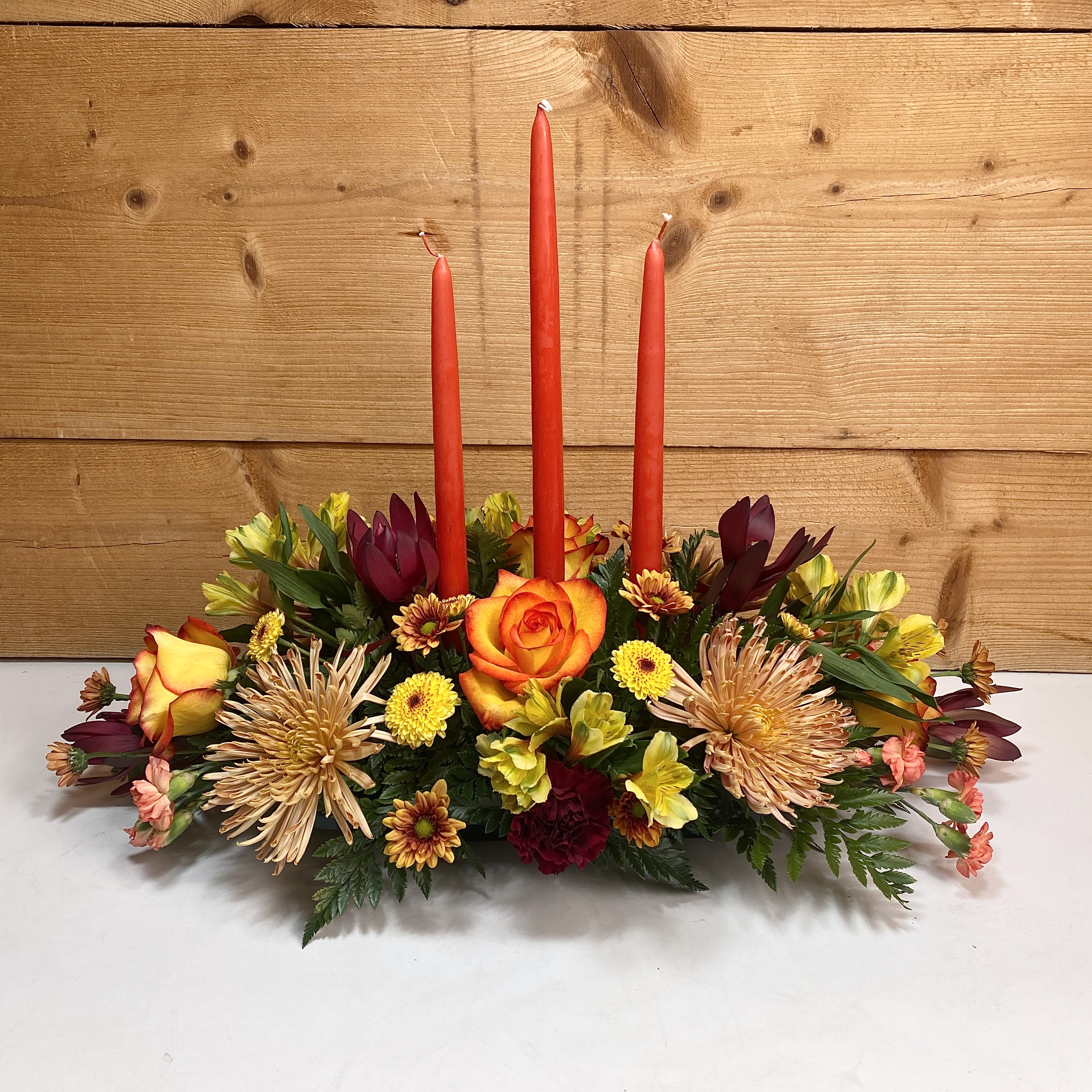 Thanksgiving Grandeur - If you're looking for a magnificent display for Thanksgiving, this arrangement is the choice for you. Thanksgiving Grandeur is a three-candle centerpiece with three, 15&quot; orange candles, and a lovely assortment of fall colored flowers including roses, alstroemeria, carnations, daisies, and more. This centerpiece would make the perfect final touch for your amazing Thanksgiving dinner, order yours today and make your Thanksgiving decorating a breeze.  Approx. 27&quot;x20&quot;
