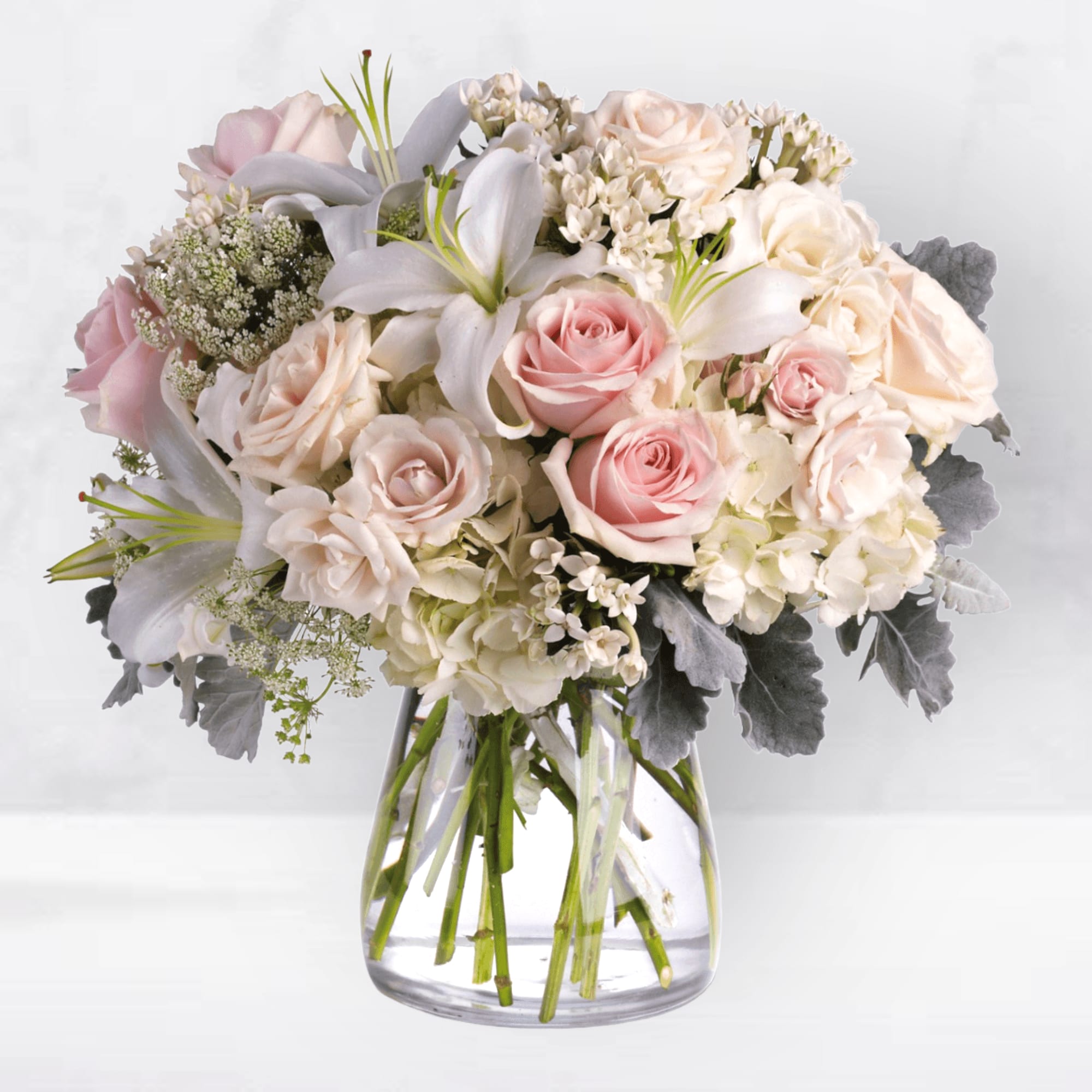 Beautiful Whisper Bouquet - The Beautiful Whisper Bouquet is an elegant floral arrangement that exudes grace and sophistication. Soft as a beautiful whisper bouquet, this elegantly feminine vase of delicate pink and white roses, hydrangea, and lilies is a simple and heartfelt affirmation of love and devotion.  White, crème, and light pink roses nestled with hydrangea, lilies, Queen Anne's lace, bouvardia and dusty miller in a classic clear-glass cinched hurricane vase. Orientation: All-Around Size: &nbsp;15 1/2&quot; H X &nbsp;15 1/2&quot; W  
