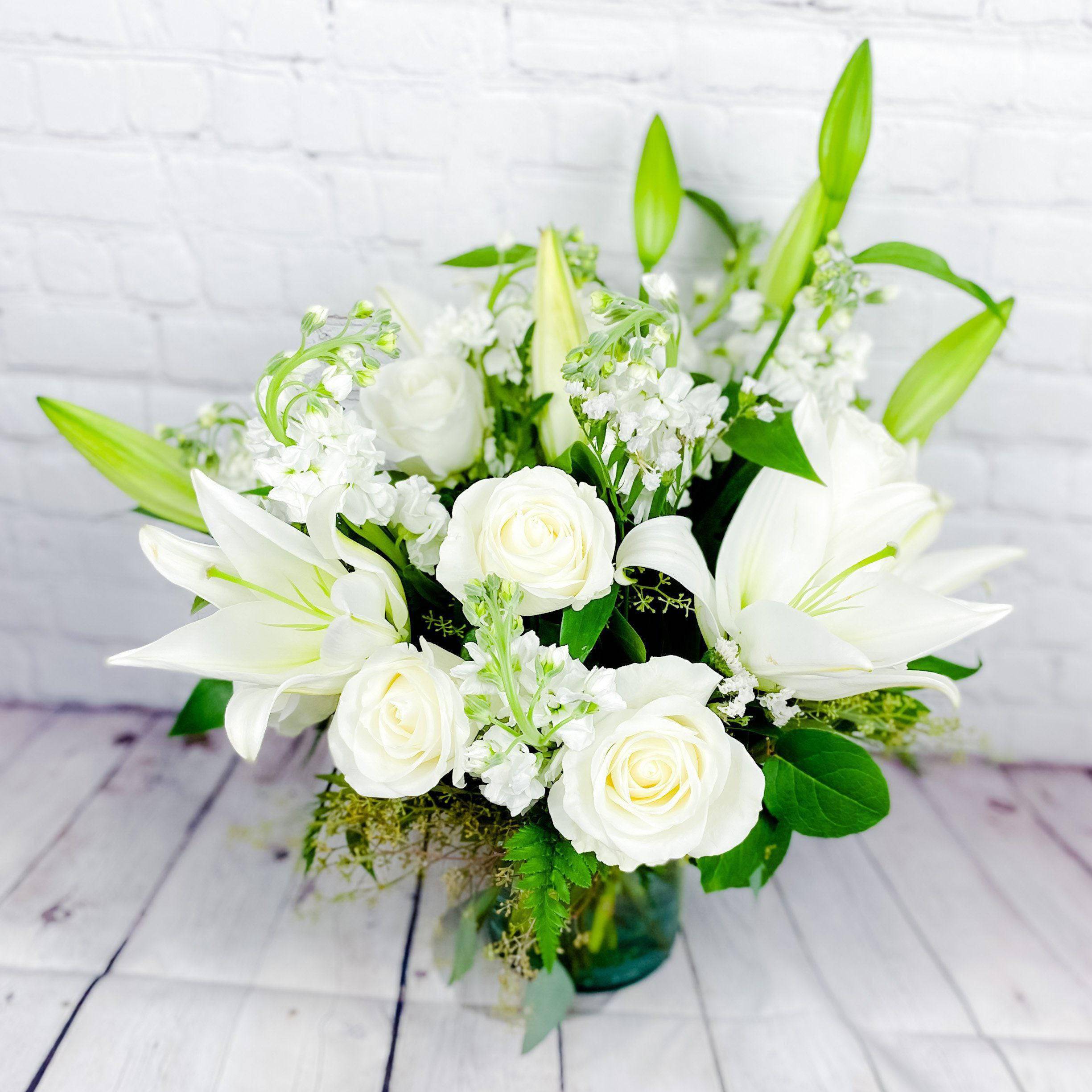 Classic All Whites By DGM Flowers - Elevate any space with the Classic All Whites by DGM Flowers, a stunning white floral arrangement. This exquisite bouquet features fresh cut flowers. Glorious white roses lead the way in this luxurious classic all whites arrangement. Paired with lilies, stocks and salal, they're hand-gathered in a stylish cylinder vase to evoke an evening stroll.  Vase measures 8&quot;H.