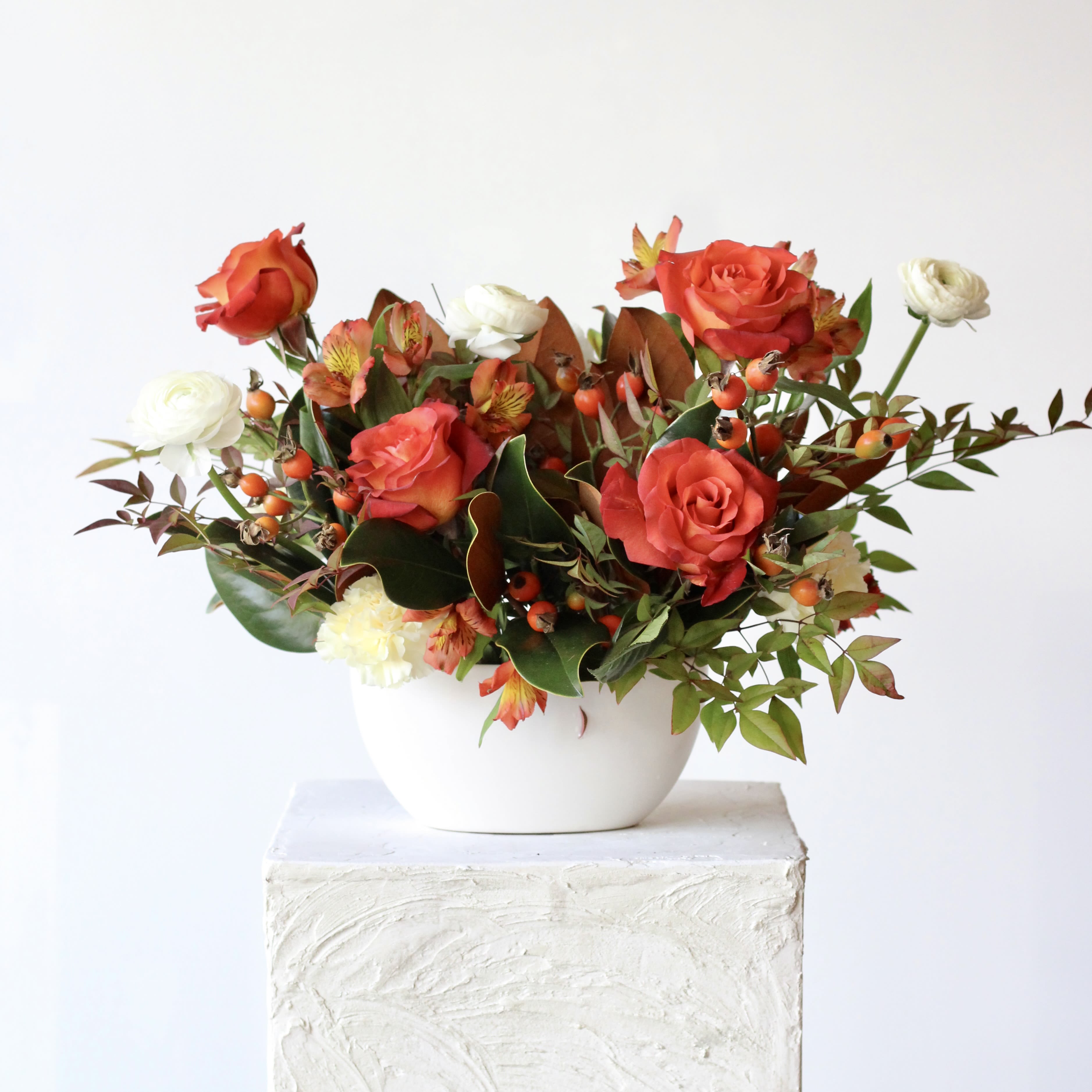 Thanksgiving Collection: Magnolia Meadows Centerpiece Thanksgiving Collection: Magnolia Meadows Centerpiece - Transform your Thanksgiving table into a masterpiece with the "Magnolia Meadows" centerpiece! Designed for hosts who want to impress, this stunning arrangement is crafted with roses, magnolia tips, and button mums. Nestled in the elegant long and low white metta boat, it offers a bold, high-contrast color palette that stands out from typical floral decor. The uniquely colored roses add a touch of sophistication and charm, making this centerpiece a conversation starter at any gathering. Celebrate the season with a burst of colors and textures that express gratitude and warmth.