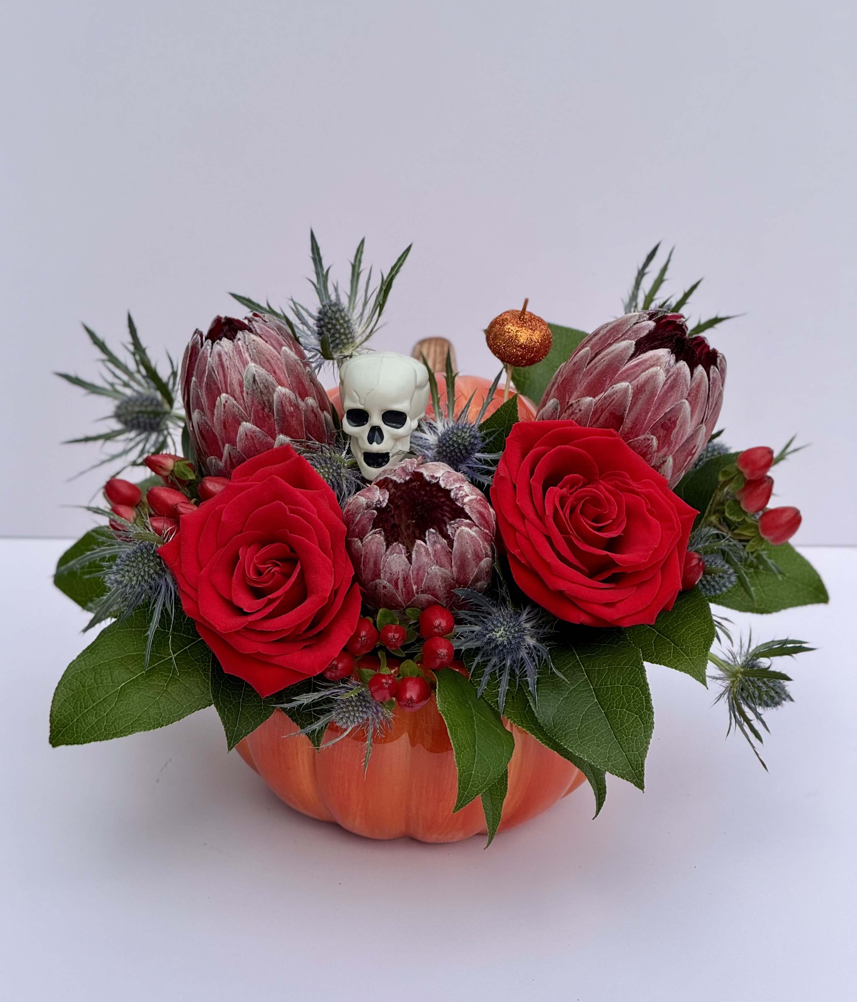 Crimson Spell - A bewitching blend of protea, roses, thistle, and autumn textures — designed in a pumpkin vessel that feels straight out of a spellbook. Each arrangement captures the warmth of fall with deep crimson blooms and moody botanical accents, perfect for Halloween tables or seasonal gifts.  Limited availability through October 31. Every piece is handcrafted and uniquely one-of-a-kind.