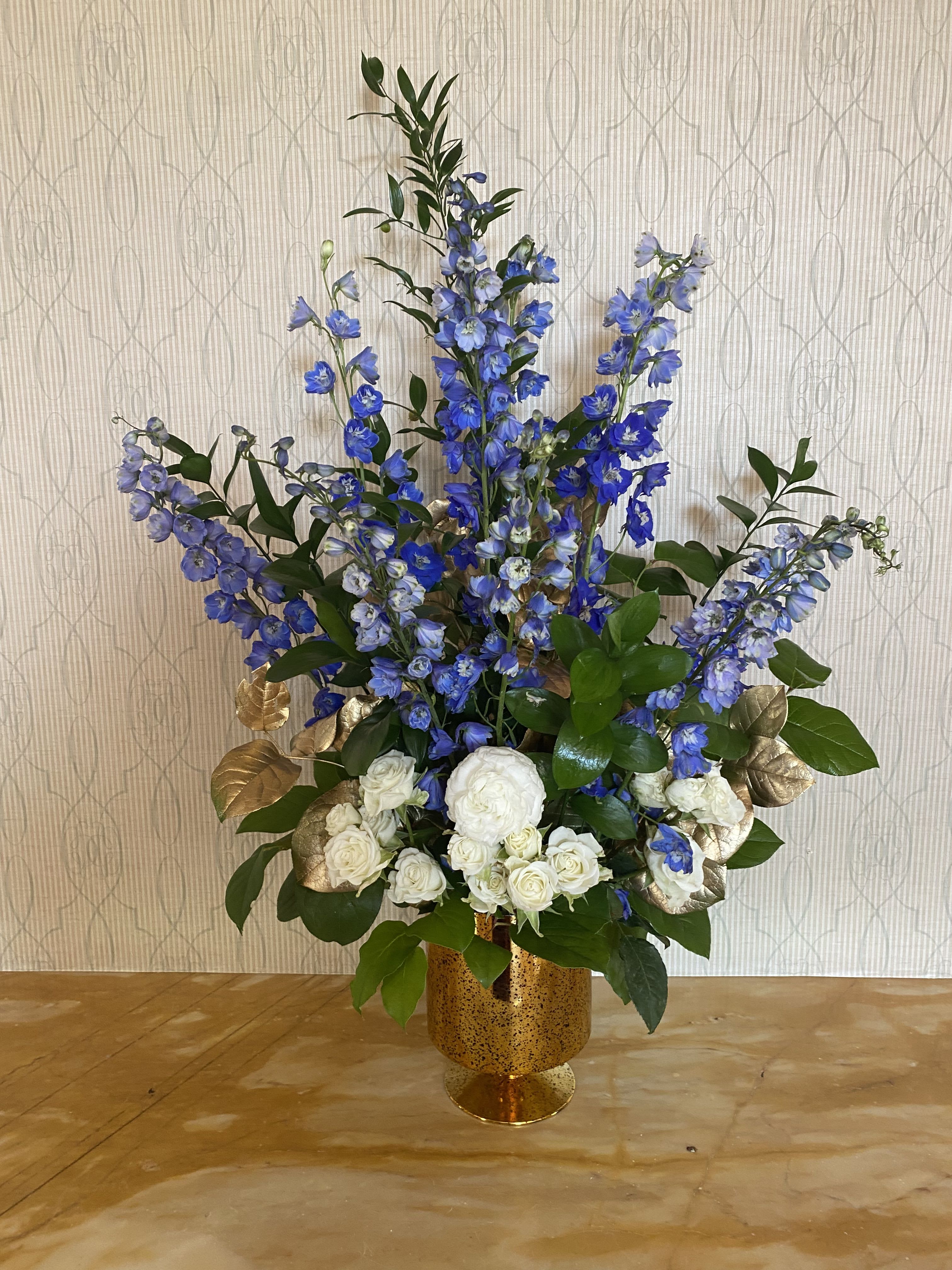 Blue and Gold Blue and Gold - A tall one sided table arrangement of blue delphinium and white roses with foliage sprayed gold in a gold mercury glass pedestal vase. Approximately 30”-36” T and 15”-24” W.