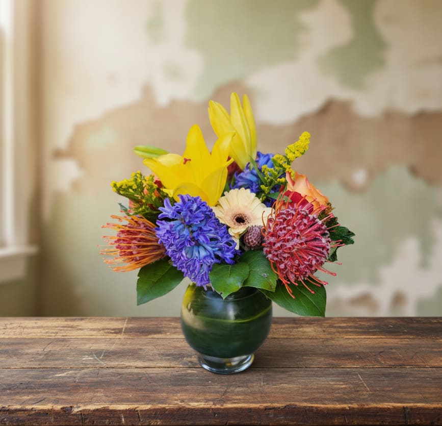 Olympia Vibrant Floral Bouquet -  Introducing our vibrant masterpiece, designed to captivate hearts and uplift spirits. This bouquet is a celebration of hues, boasting vibrant yellow lilies, delicate blue hyacinths, and striking red protea. Each flower is carefully selected to create a harmonious symphony of colors that will leave a lasting impression.  The design style of this bouquet is compact yet impactful, ensuring it packs a punch wherever it's placed. Whether adorning a work desk or gracing an end table in a bedroom, this bouquet adds a touch of sophistication to any space.  Perfect for occasions like Mother's Day, birthdays, or to convey heartfelt wishes for a speedy recovery, this bouquet is guaranteed to brighten someone's day. It's a thoughtful gesture that speaks volumes, conveying warmth and affection with every bloom.  Please note that while some flowers may be seasonal, Specialty Floral Design is committed to ensuring your bouquet maintains its essence. We'll make suitable substitutes to ensure that the look and feel of your arrangement remains intact, ensuring every recipient receives a stunning display of nature's beauty.   Specialty Floral Design is more than just a local florist; we're your partners in crafting unforgettable moments and meaningful connections. Serving Olympia, WA, Tumwater, WA, and Lacey, WA, as well as surrounding areas in Thurston County to Lewis County, we're committed to bringing joy and beauty to your doorstep. 