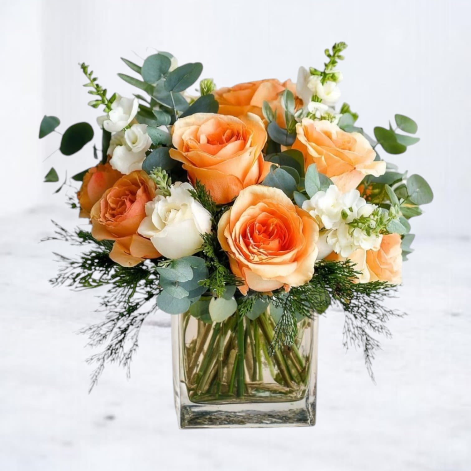 Apricot Candor - The Apricot Candor bouquet is a glowing blend of cheerful charm and timeless elegance. Featuring vibrant orange roses, creamy white spray roses, and delicate stock, accented with lush eucalyptus, this arrangement brings a sense of warmth and joy to any occasion. Designed in a clear glass vase, it’s a versatile gift that’s perfect for birthdays, get well, or simply brightening someone’s day. Hand-delivered with care, this bouquet radiates beauty and sophistication, making it a heartfelt way to celebrate life’s special moments.