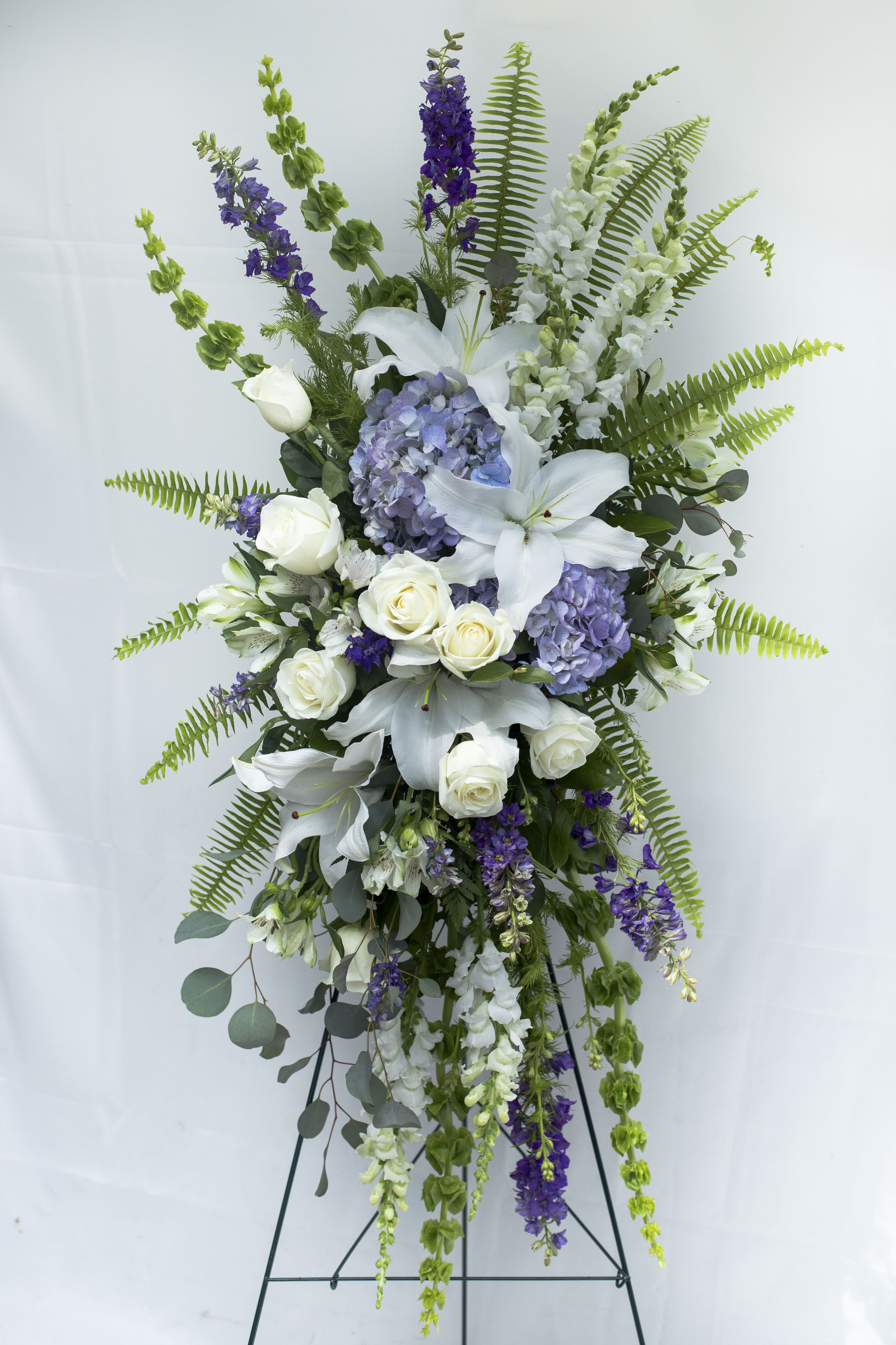 Always Loved Standing Spray - What better way to express your love then through this tribute of blue Hydrangea, white Lilies, white Roses, purple Larkspur, Bells of Ireland, and accented with Sword Fern greenery. This spray is stately in the length of approximately 48&quot; and 30&quot; Wide.