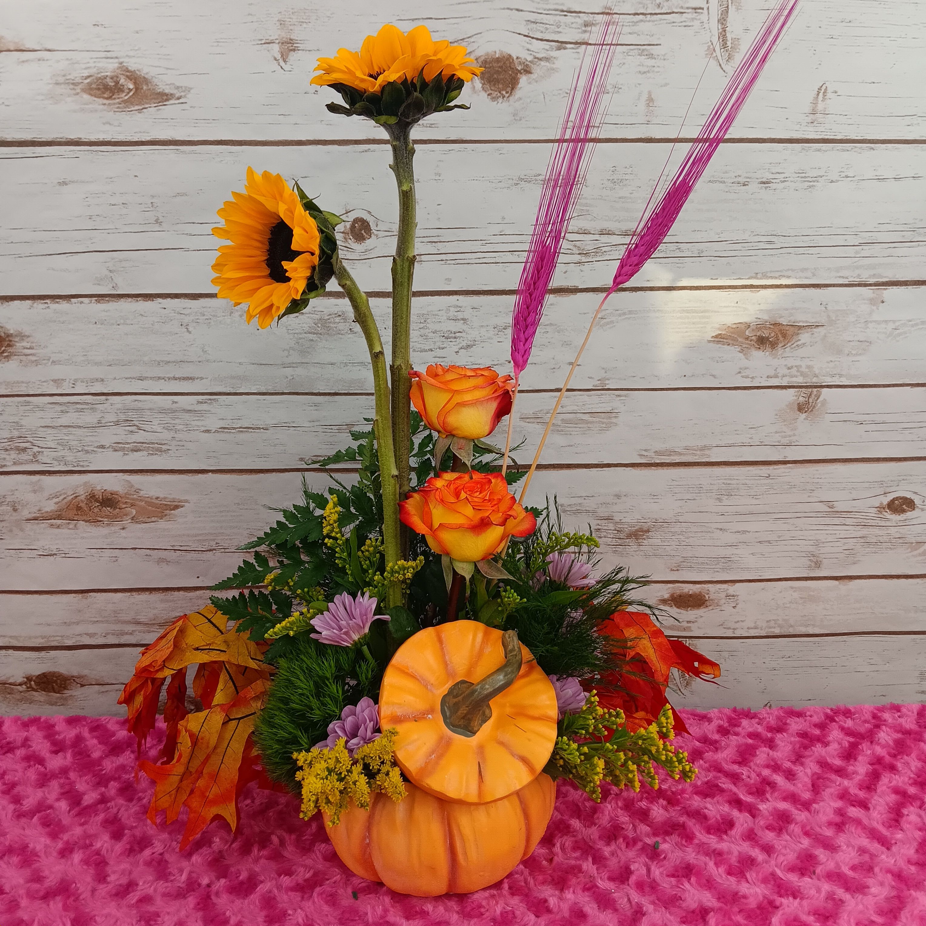 SUNFLOWER PUMPKIN - Bring the warmth of autumn indoors with this cheerful pumpkin-inspired arrangement. Bright golden sunflowers take center stage, beautifully complemented by roses, blooms, and accents of fresh greenery. Designed in a festive ceramic pumpkin vase, this arrangement is perfect for celebrating the season, adding a touch of fall charm to your home, or gifting to someone special. Disclaimer: Sunflower size may vary due to seasonal changes.