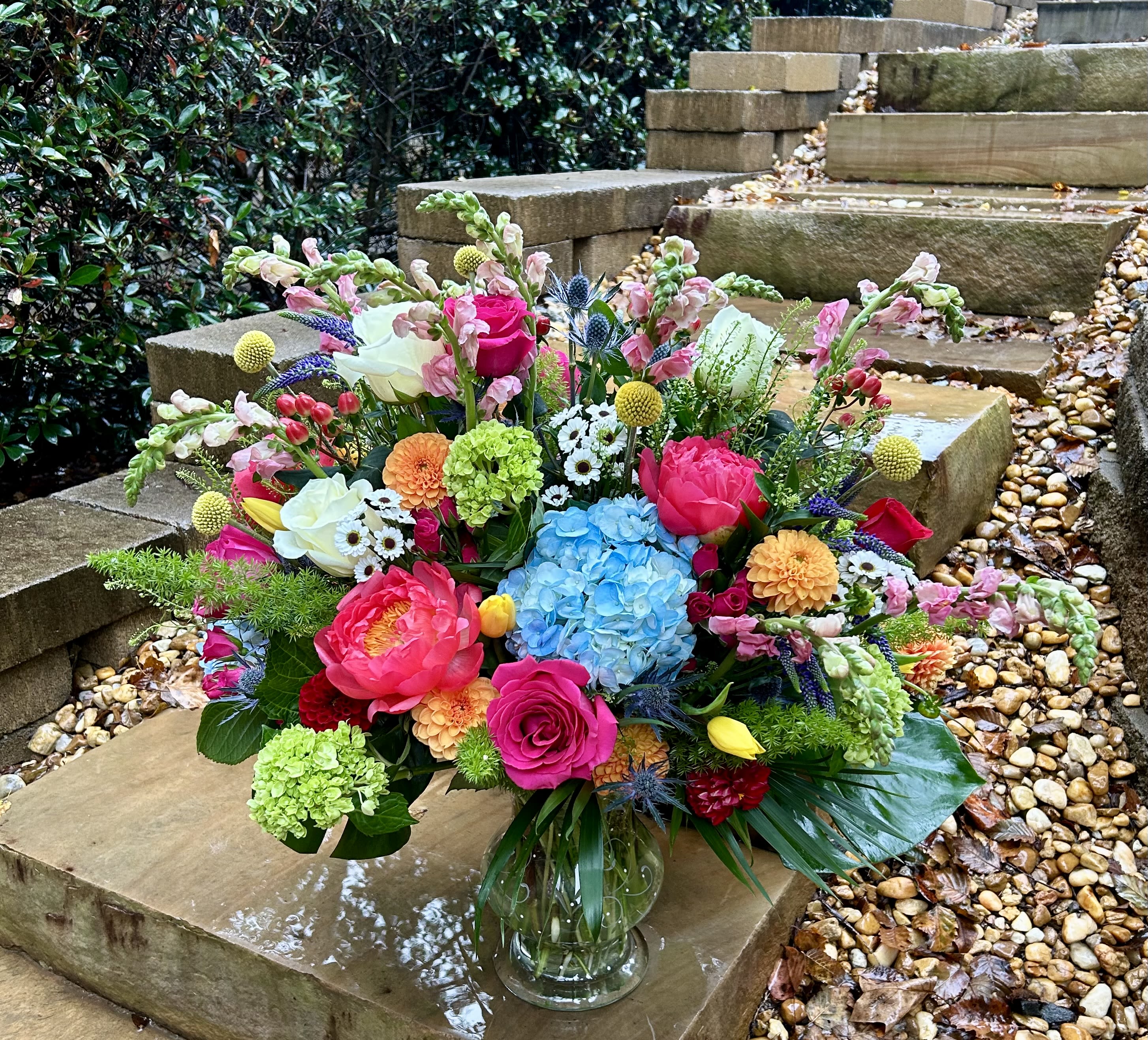 Garden Jubilee - A lavish garden-inspired creation bursting with color and texture — from blue hydrangeas and coral peonies to golden craspedia, white daisies, and verdant greens. Set in a clear vase, this arrangement captures the exuberance and abundance of a summer garden in full bloom.