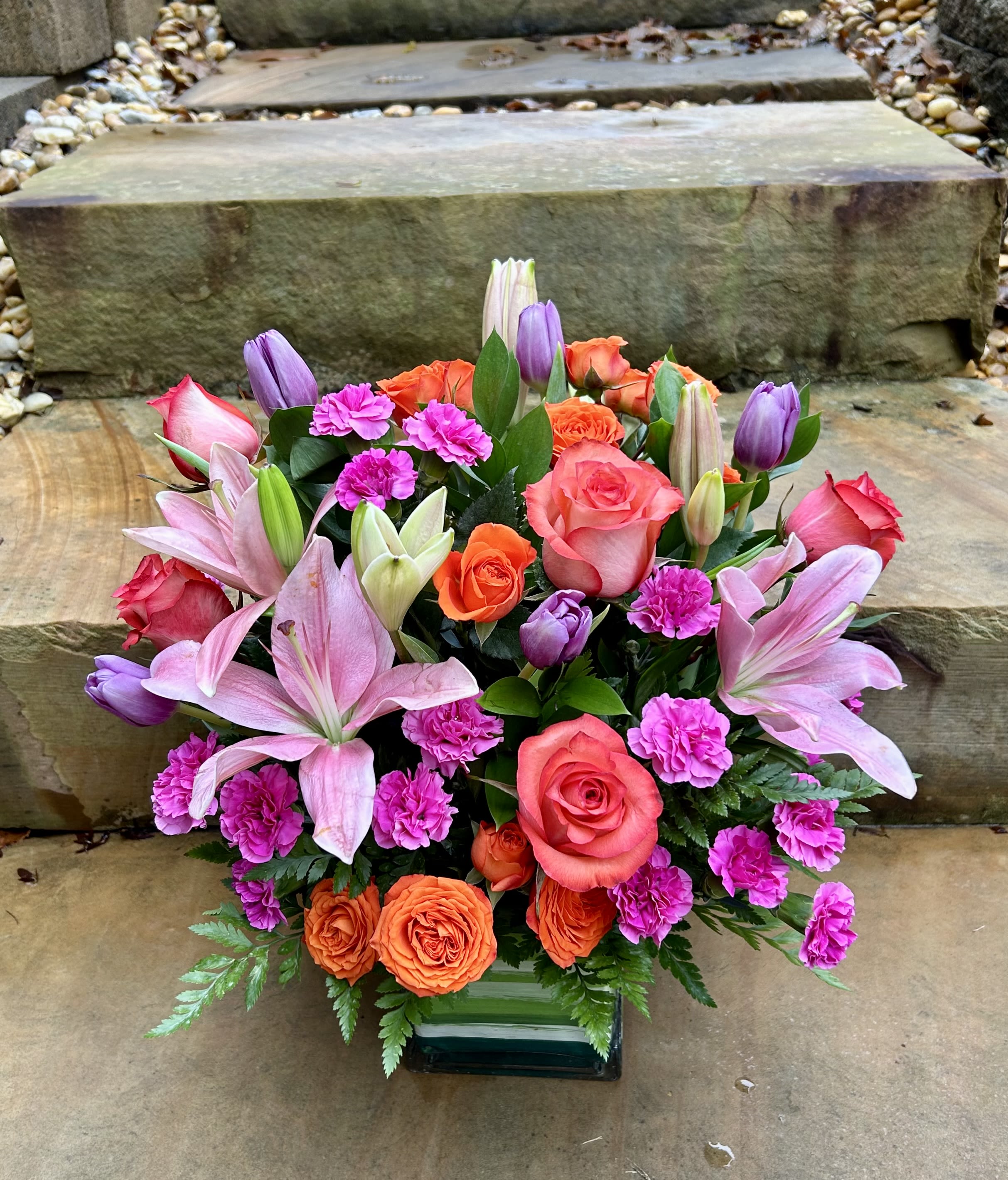 Sunset Harmony - A bold symphony of color featuring pink lilies, coral roses, orange spray roses, fuchsia carnations, and touches of lavender tulips. Arranged in a sleek square vase, this design balances elegance and energy — perfect for brightening any room or celebration. 