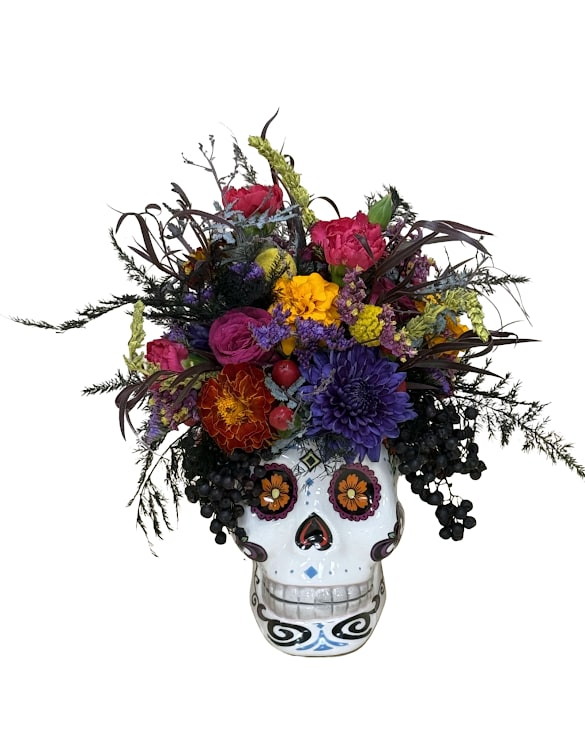 Bloom of the Dead - Celebrate Día de los Muertos with this stunning one-sided floral display, artfully arranged in a keepsake ceramic sugar skull container. Featuring a rich mix of marigolds, roses, and accent blooms in vibrant shades of gold, purple, and fuchsia, this design captures the beauty and meaning of remembrance. Perfect as a centerpiece or tribute piece, it brings color, culture, and heart to any celebration of life.  Approximate size: 8.5”H x 8”W.