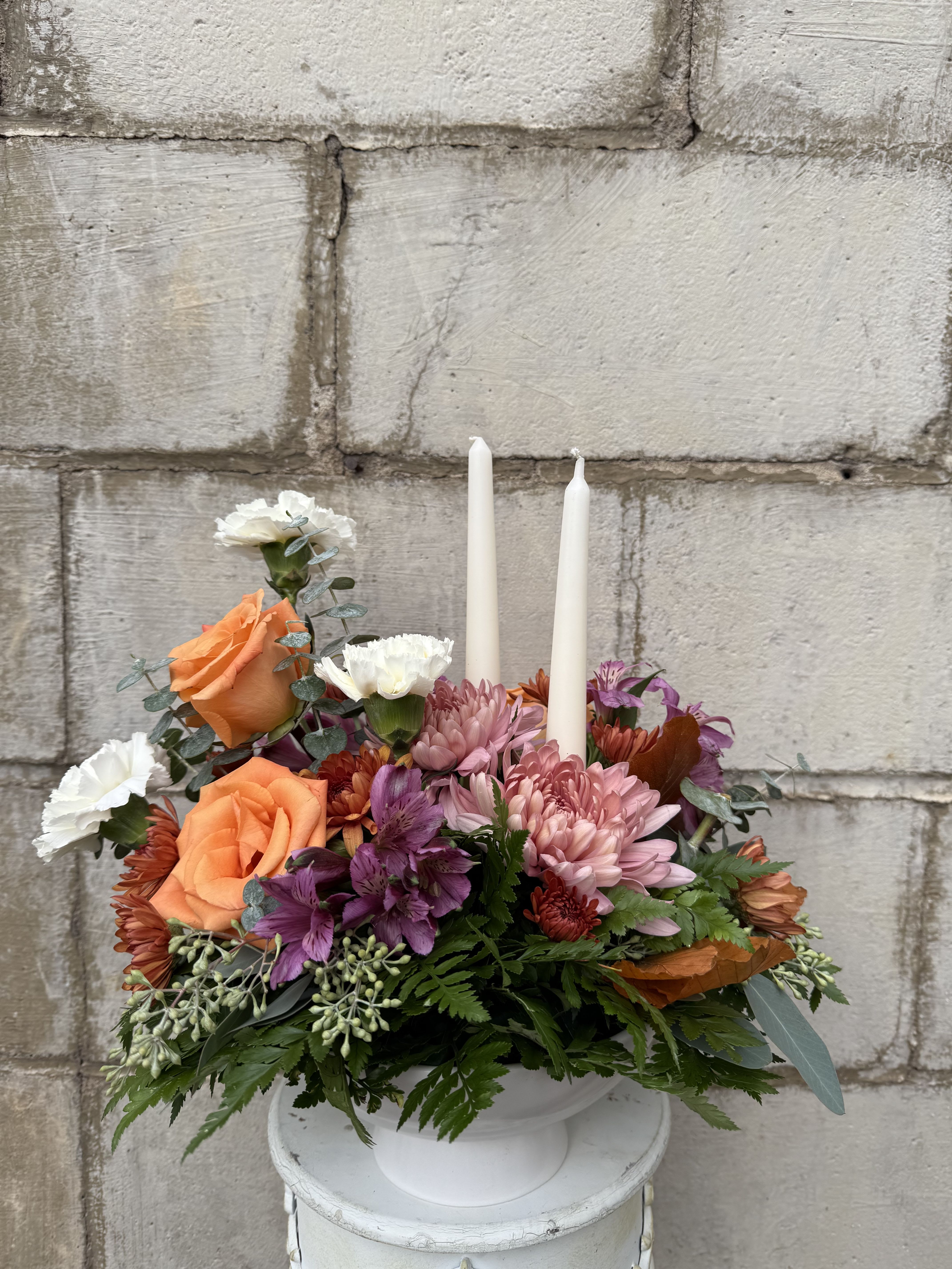 Autumn Grace Centerpiece - Celebrate the beauty of the season with our Autumn Grace Centerpiece — an elegant Thanksgiving table arrangement designed to bring warmth and charm to your holiday gathering. Nestled in a classic white footed ceramic compote, this arrangement features vibrant orange roses, delicate white carnations, purple alstroemeria, peach cremones, and bronze cushion mums, all surrounded by lush greenery. Two graceful white taper candles add a soft, romantic glow, creating the perfect ambiance for sharing gratitude, good food, and heartfelt moments around the table.  All flowers, flower colors, and containers are subject to change due to availability. 