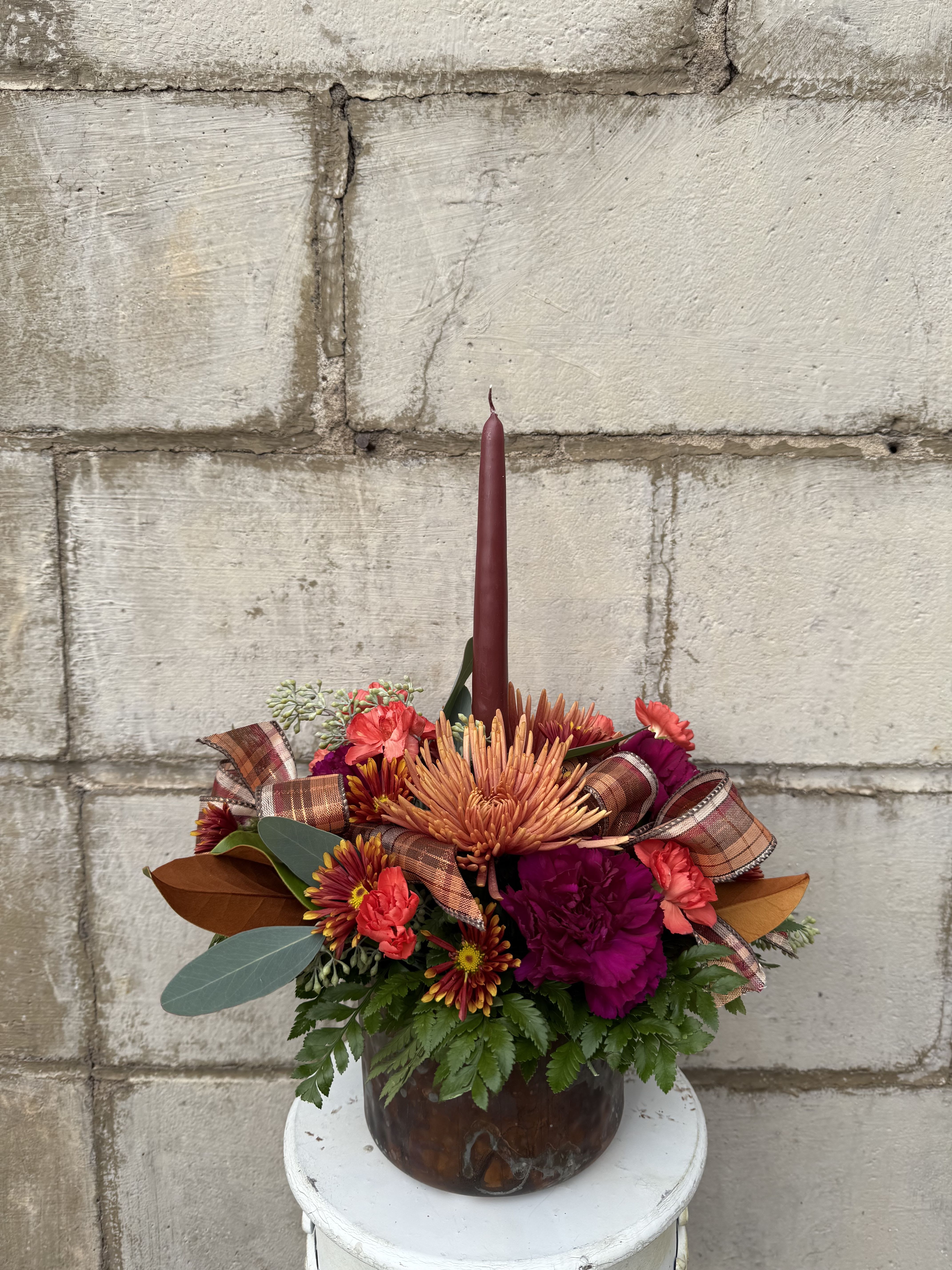 Copper &amp; Cranberry Centerpiece - Warm, rich, and full of fall charm — the Copper &amp; Cranberry Centerpiece captures the essence of Thanksgiving in every detail. Designed in a metal copper pot, this arrangement features a lush mix of bicolor and bronze cushion mums, orange mini carnations, and purple carnations, accented with seasonal greenery and a coordinating ribbon. A single burgundy taper candle adds a soft, cranberry-toned glow, creating the perfect cozy focal point for your holiday table or home décor.  All flowers, flower colors, and containers are subject to change due to availability.