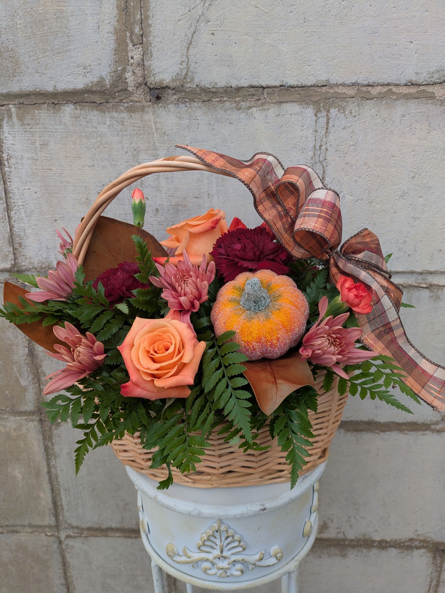 Golden Gatherings Centerpiece - Celebrate the heart of the season with our Golden Gatherings Centerpiece — a radiant arrangement designed to bring warmth and beauty to your Thanksgiving table. Nestled in a natural harvest basket, this piece features orange roses, burgundy carnations, orange mini carnations, and salmon cushion mums, accented with lush greenery, a decorative pumpkin, and a seasonal ribbon. Bursting with golden autumn tones, it’s the perfect way to add a welcoming, festive touch to your holiday gatherings and moments of gratitude.  All flowers, flower colors, and containers are subject to change due to availability.