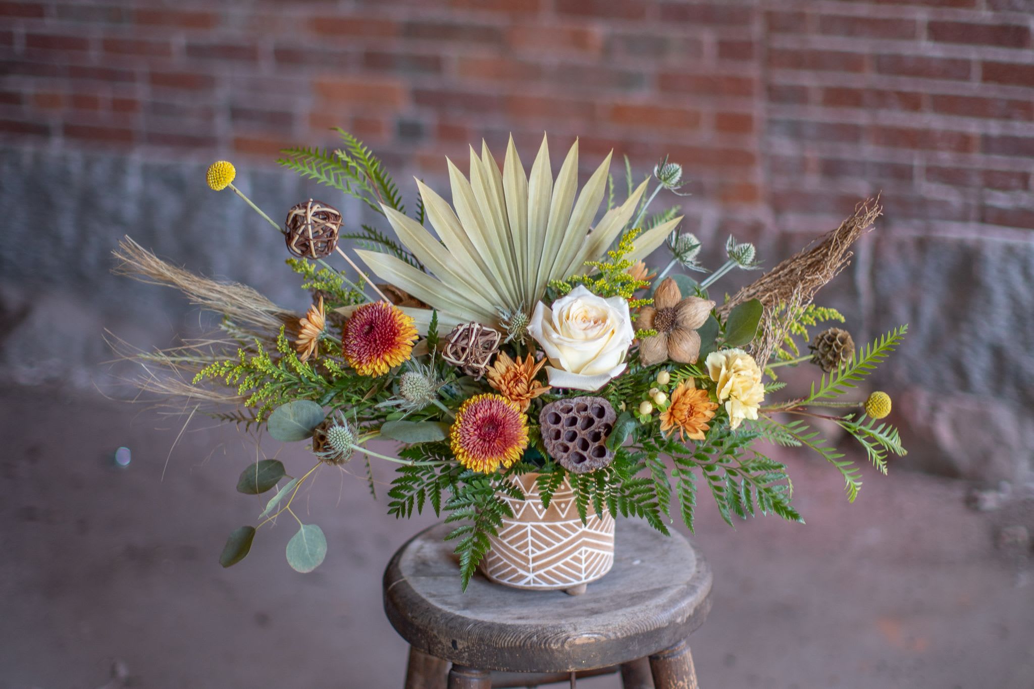 Texture FANatic - A perfect medley of fresh and dried floral materials artfully arranged in an elegant matte finish ceramic planter. An exotic preserved palm fan is the focal point of this unique design. Encompassing the palm are textural elements galore, both fresh and dried. Thistle, scabiosa, craspedia, lotus pods and natural wheat dance with fresh fall pom pons, chrysanthemums and a pastel tone rose. Bloom colors may vary but a muted fall palette will be custom curated based on our freshest inventory. 