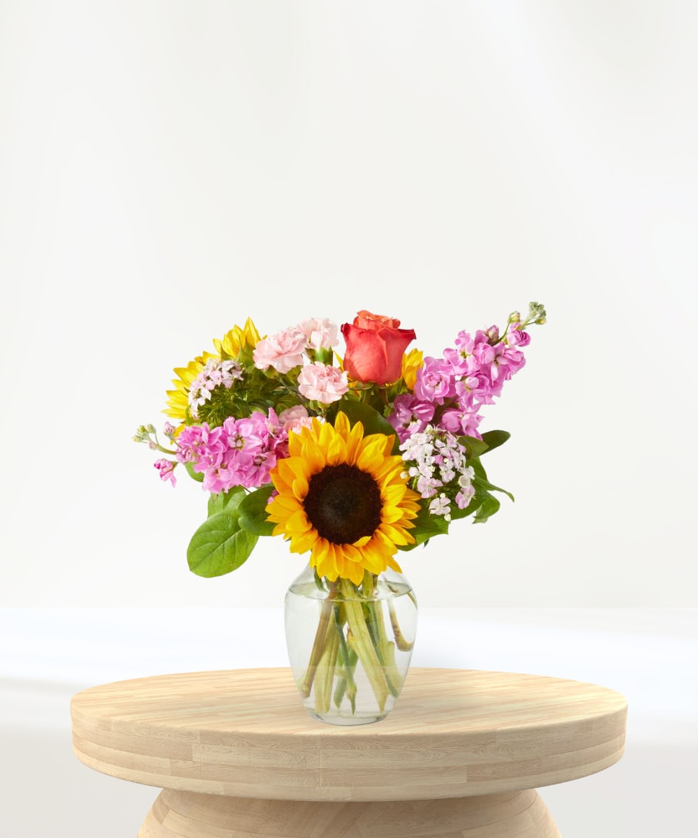 Sun-Kissed Garden - Brighten their day with this cheerful burst of color! Featuring a vibrant mix of golden sunflowers, soft pink stock, fragrant carnations, lavender blossoms, and a touch of classic orange rose charm, this hand-crafted arrangement brings joy and beauty to any space. Artfully arranged in a clear glass vase, it’s perfect for birthdays, celebrations, thank-you's, or just because.  What’s included: Sunflowers, Pink Stock, Carnations, Orange Roses, and Seasonal Foliage Arranged in a clear glass vase