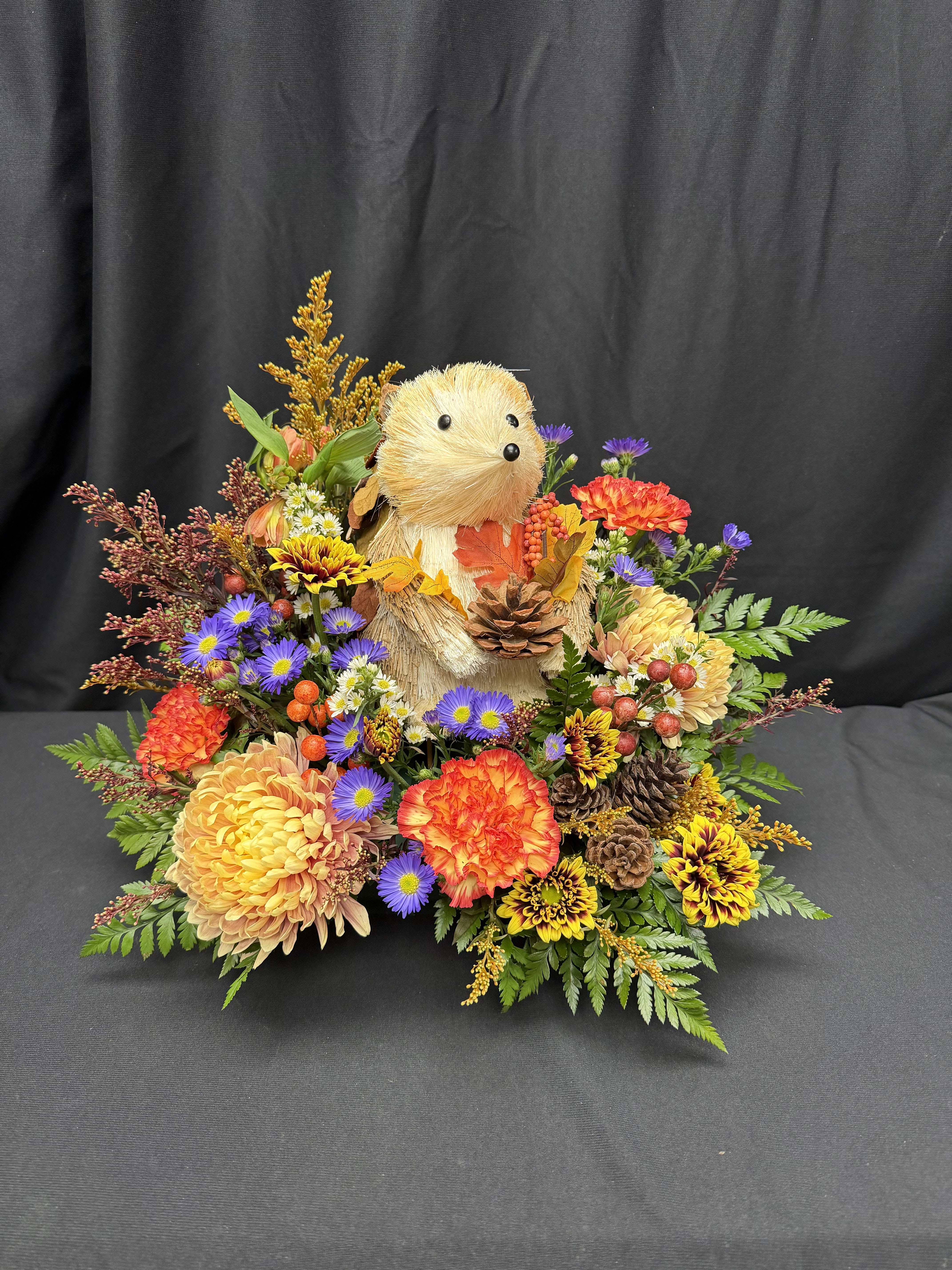 Hedgehog Harvest - Celebrate the cozy charm of autumn with our delightful Hedgehog Harvest arrangement! Featuring a whimsical hedgehog keepsake nestled among vibrant fall blooms, this design bursts with warm tones of orange, gold, and plum. Accented with pinecones, berries, and seasonal textures, it’s the perfect way to bring a touch of woodland magic to your home or gift to someone special.  Includes a reusable hedgehog keepsake — a sweet reminder of the season’s joy long after the flowers have faded.
