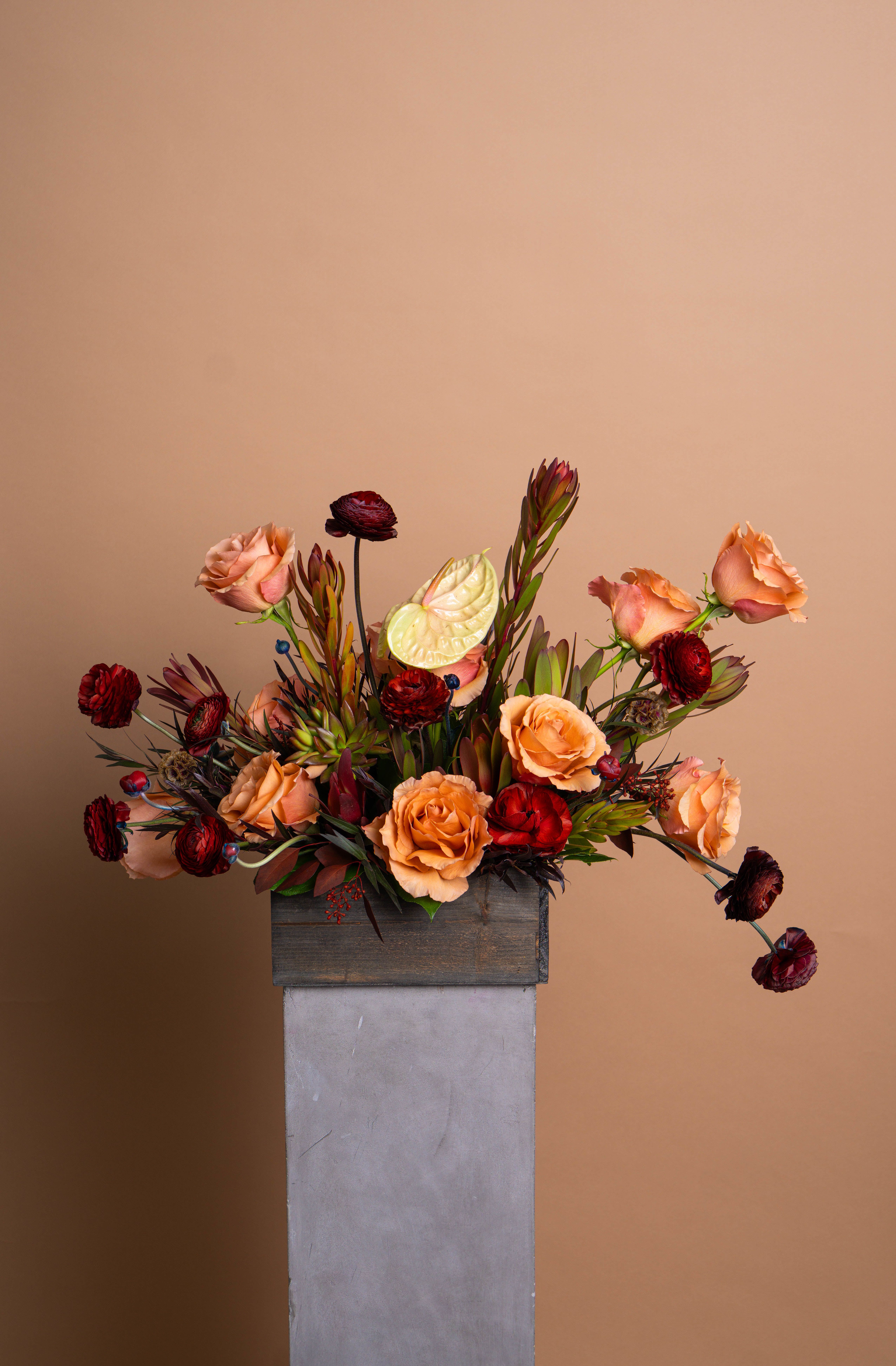 HOPE - Warm and sculptural, Hope blends sunset tones with quiet intensity. Amber and peach roses meet deep burgundy ranunculus and textural leucadendron, anchored by soft greenery and a single pale anthurium for contrast. The result is both modern and timeless, a composition that radiates understated confidence.  Refined, rich, and full of movement, Hope evokes strength wrapped in elegance.  The arrangement pictured is Deluxe.
