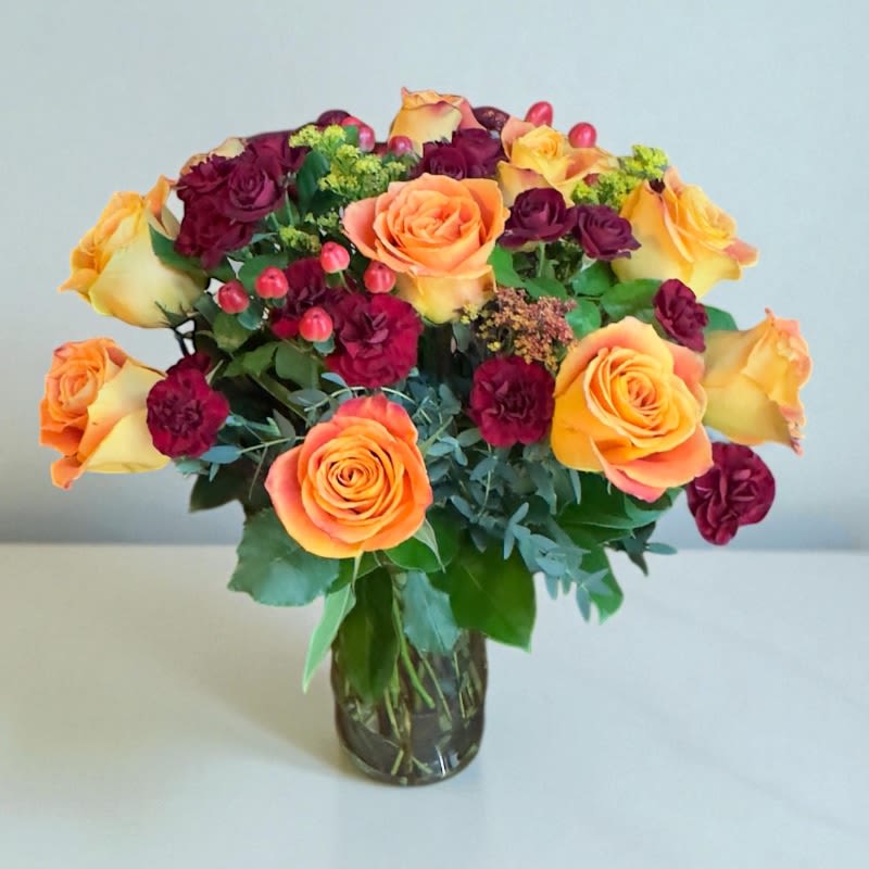 Golden Ember #82 - Warm, vibrant, and full of autumn charm — Golden Ember is the perfect gift to express gratitude, love, or simply brighten someone’s day. This radiant bouquet features glowing bicolored orange/yellow roses paired with deep burgundy spray roses and carnations and accented with red hypericum berries and lush seasonal greens. Each bloom brings the cozy magic of fall to life, creating a heartfelt and elegant surprise.  Beautifully arranged in a clear glass vase, Golden Ember makes a stunning centerpiece or thoughtful gift for any occasion — from birthdays and anniversaries to “just because.”   Send the warmth of autumn straight to their doorstep — order Golden Ember today and let your gesture speak in colors of love and joy!