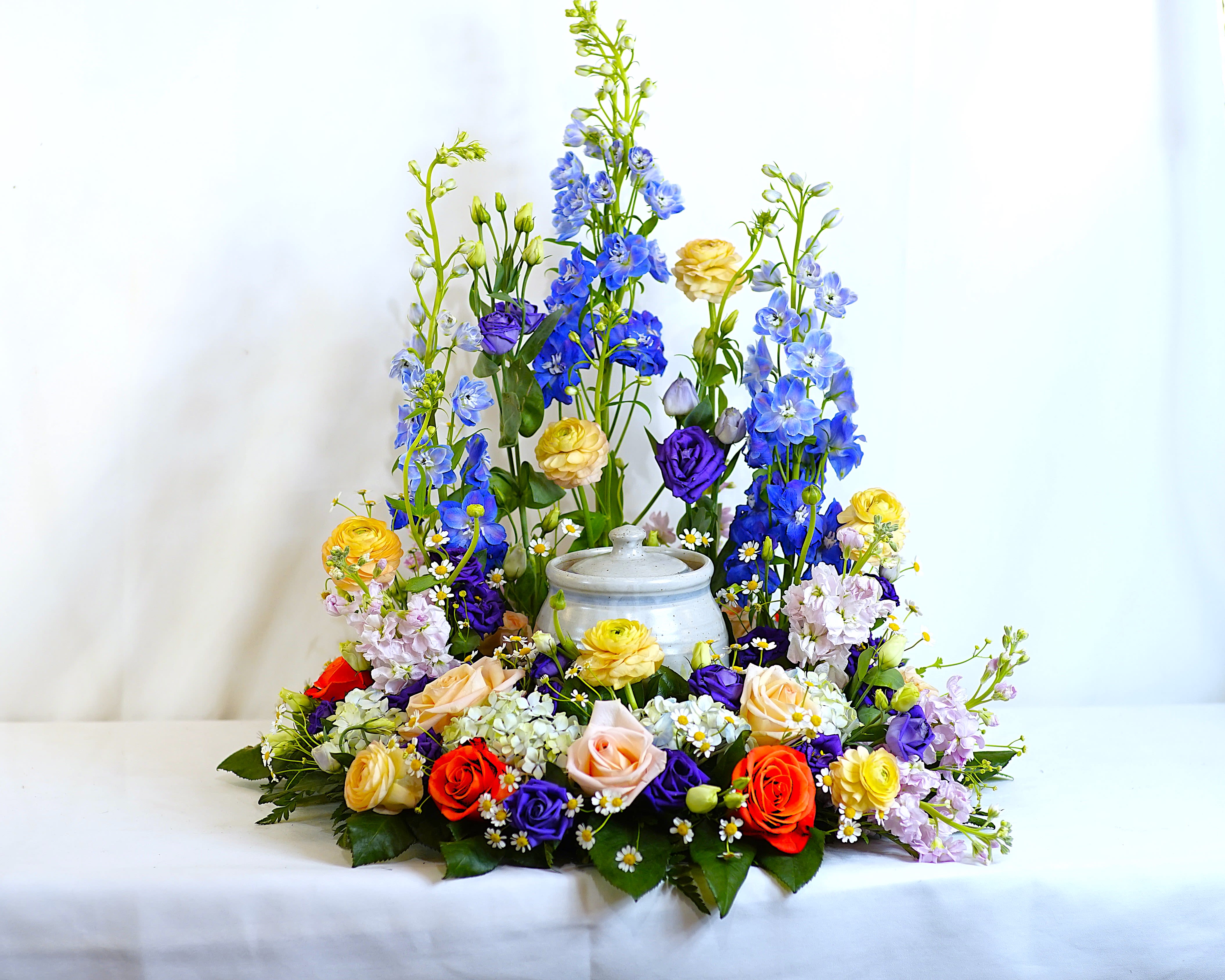 Twilight Garden Urn Tribute - A serene and elegant display, the Twilight Garden Urn Tribute is designed to surround the urn with beauty, peace, and reverence. Featuring soft lavender roses, delicate blue delphinium, regal purple lisianthus, creamy white carnations, and blush chrysanthemums, this arrangement blends cool and calming tones with lush greenery for a dignified, heartfelt tribute.  Perfect for memorial services or celebration of life ceremonies, this design brings gentle comfort and grace to any setting. Custom color palettes are available upon request to reflect your loved one’s personality or favorite blooms, ensuring a truly personal expression of remembrance.