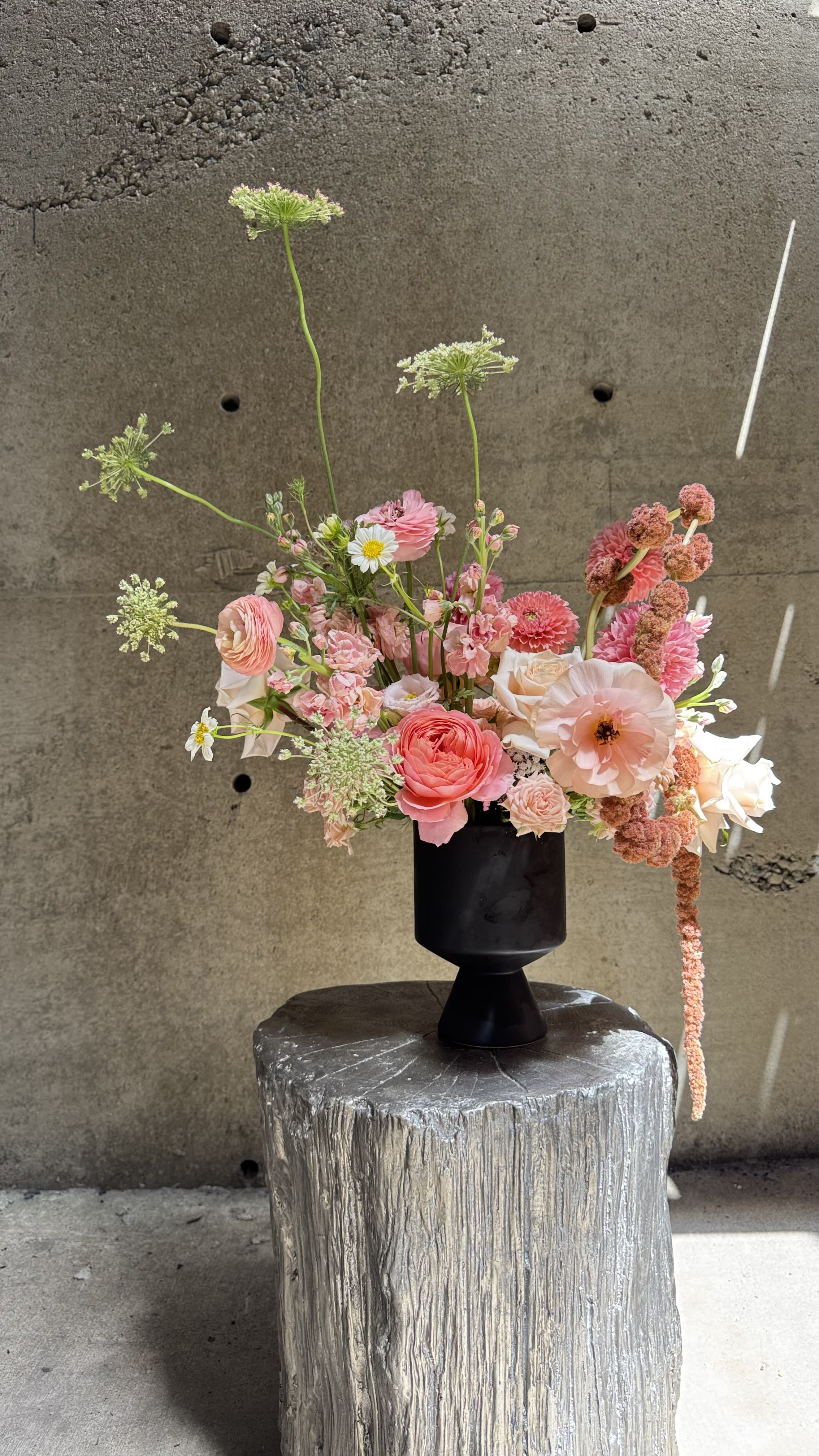 Rosé Reverie - Playful yet sophisticated, Rosé Reverie is a celebration of soft romance and modern artistry. A lush gathering of peach ranunculus, blush roses, pink dahlias, and delicate spray carnations is accented with airy Queen Anne’s lace and whimsical textures. Styled in a sleek black pedestal vase, the design balances elegance with bold contrast, making it an eye-catching centerpiece. Perfect for birthdays, anniversaries, or any occasion that calls for a touch of joy, charm, and contemporary floral beauty.