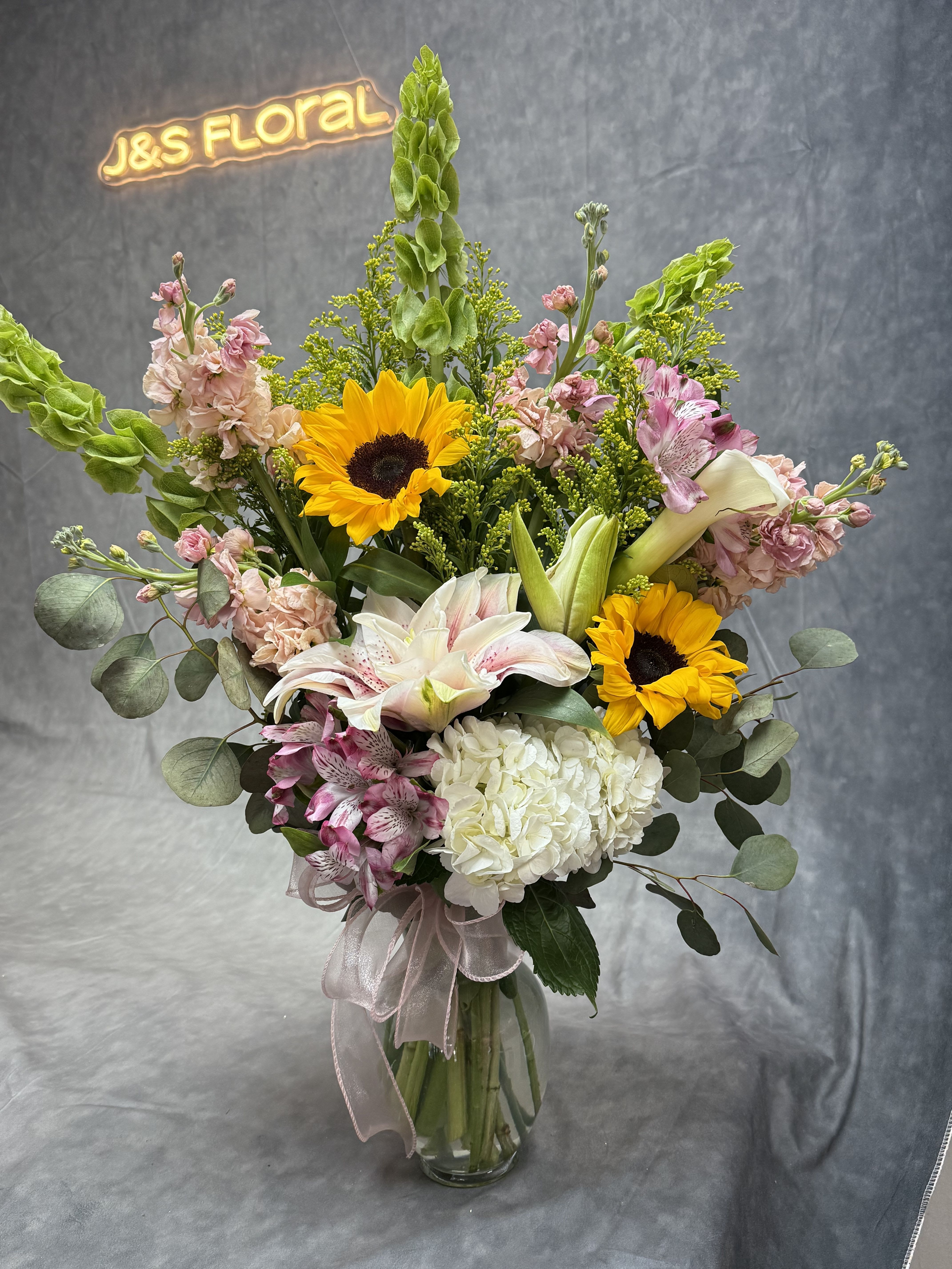 Golden Garden Symphony  - A radiant burst of elegance featuring cheerful sunflowers, lush hydrangeas, graceful lilies, striking calla lilies, Bells of Ireland, and seasonal blooms—all artfully arranged in a designer vase. This vibrant creation brings the beauty of a blooming garden right into your space. 