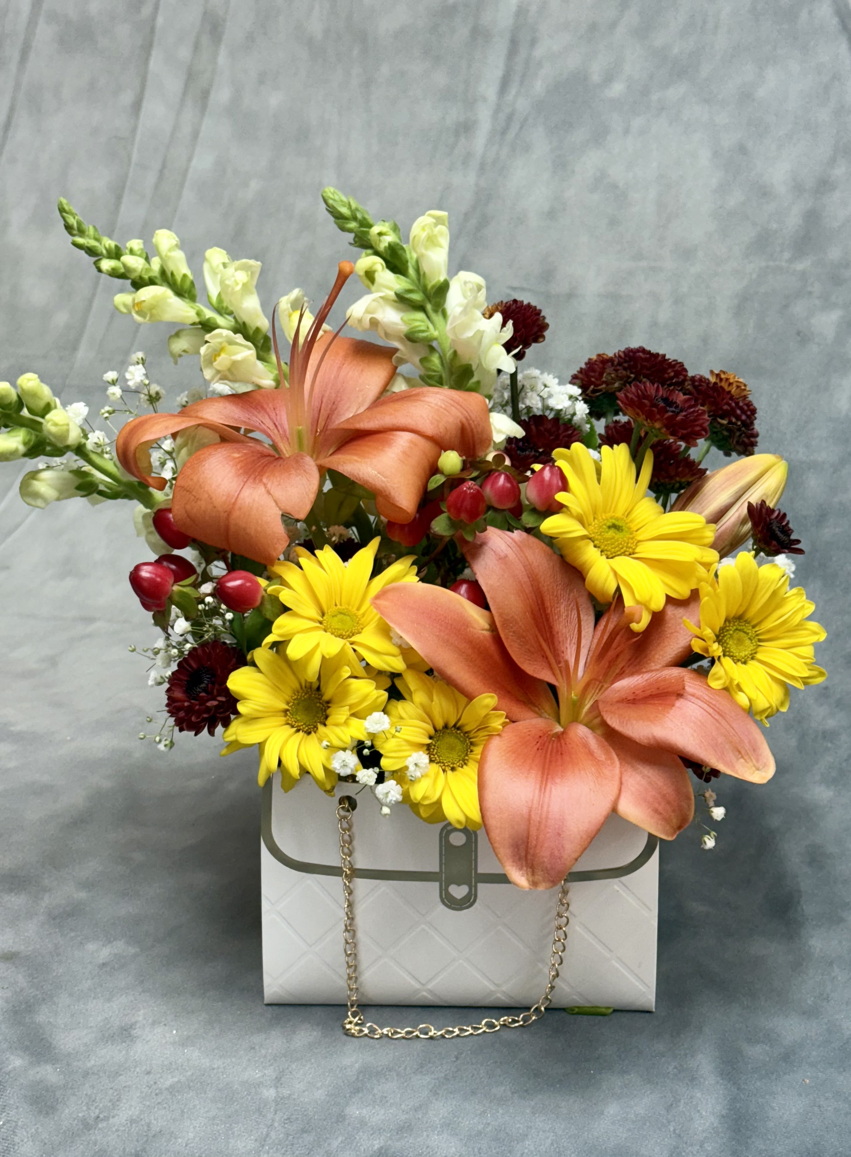 Harvest Glow Purse - Harvest Glow Purse Bring the warmth of autumn into any space with this charming harvest-inspired purse. Bursting with vibrant orange lilies, cheerful yellow daisies, and elegant snapdragons, it’s the perfect blend of seasonal beauty and rustic charm.   Upgrade Option: Make it extra cozy with our deluxe size, which includes a fall-scented candle to fill the air with the comforting aroma of the season.