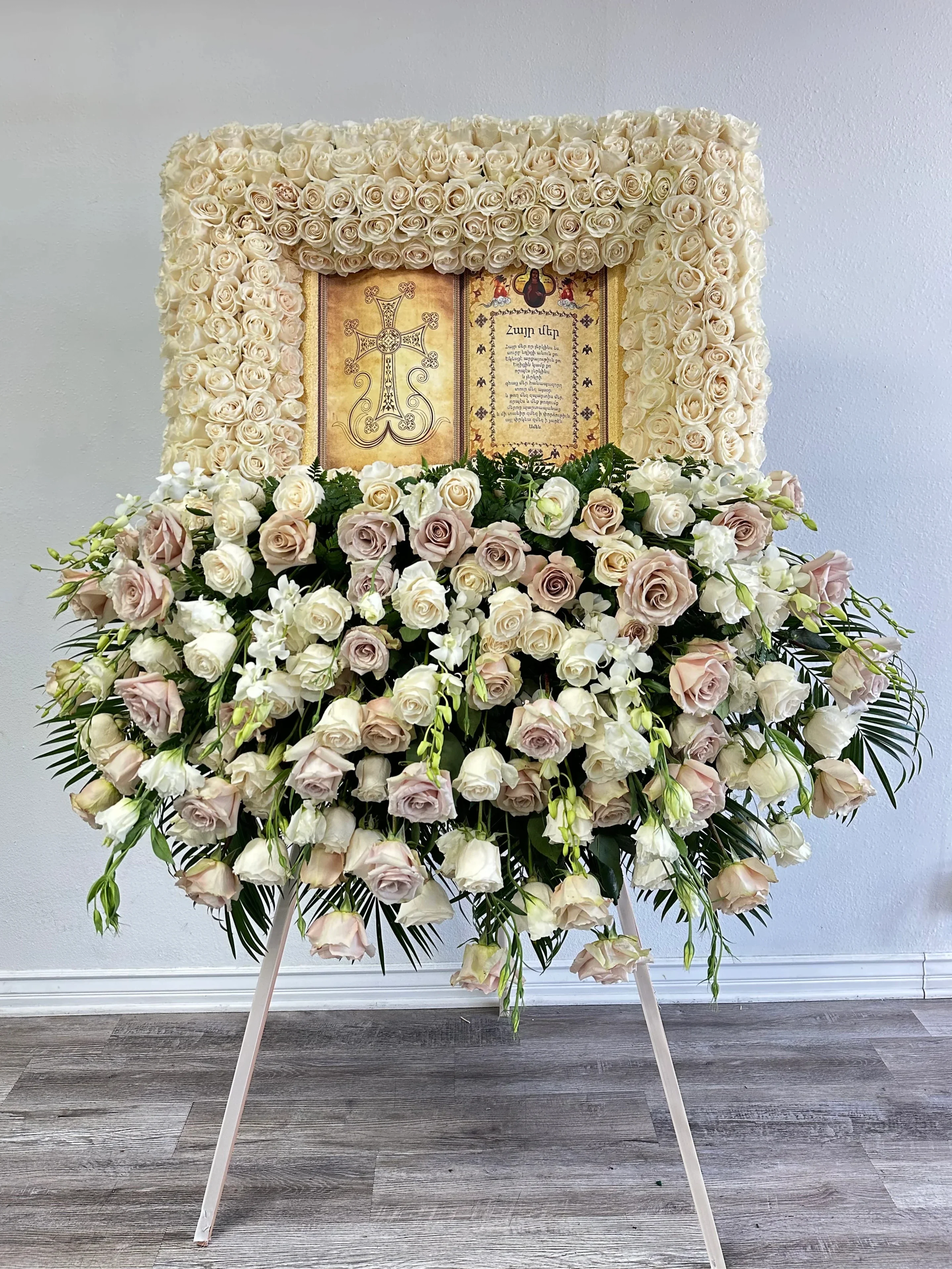 Bible Tribute with Arrangement - A luxury standing tribute featuring a framed arrangement of ivory roses surrounding a religious scripture panel, paired with a cascading display of cream and blush roses, snapdragons, and lush greenery. This design combines artistry and reverence, making it a powerful expression of faith, remembrance, and eternal love. Ideal for funeral services, memorial ceremonies, and religious tributes.