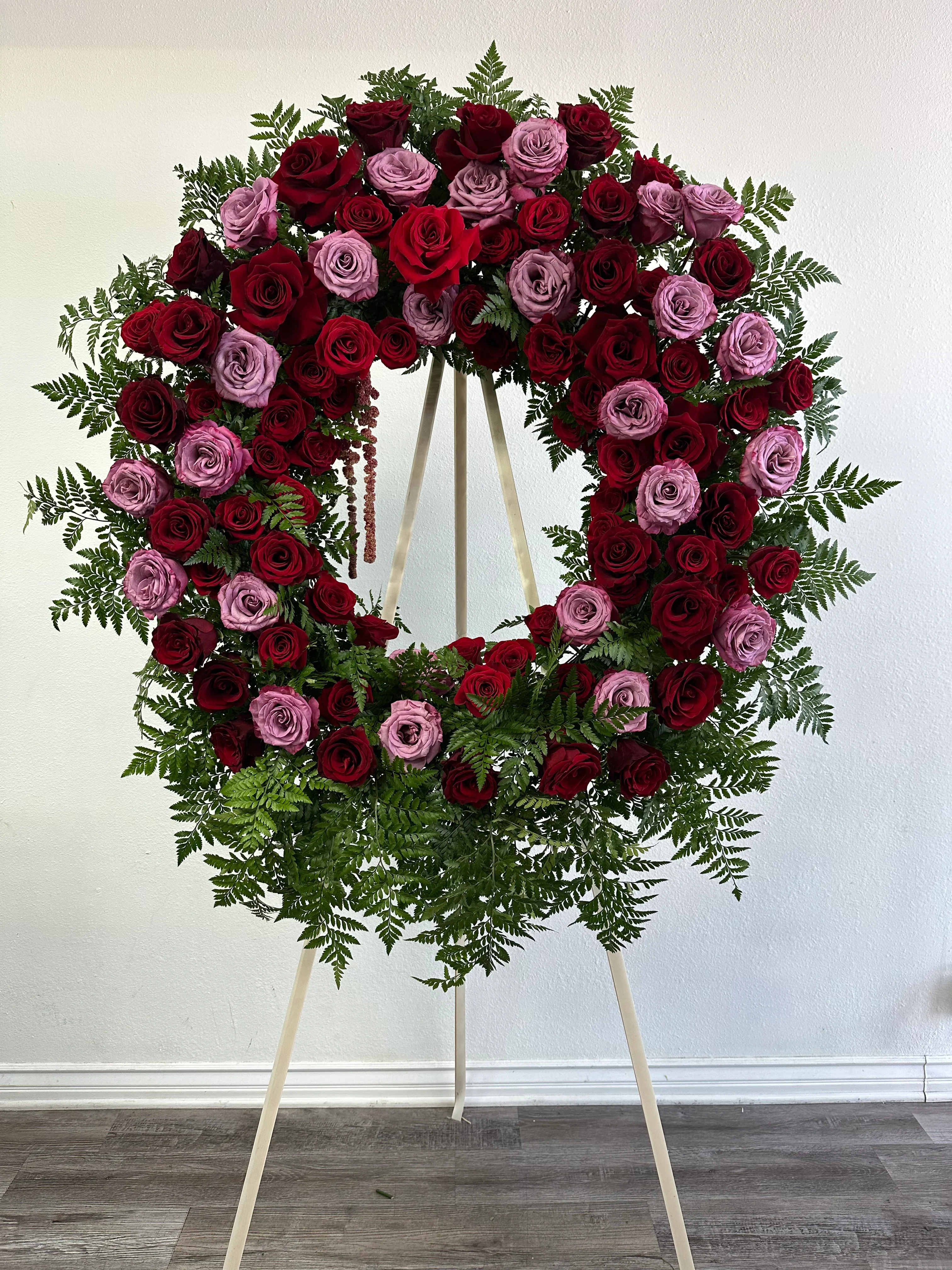 Forever in Our Hearts Wreath - A beautiful funeral/memorial wreath crafted with a rich mix of deep red and lavender roses, accented with lush greenery and ferns. Designed on a standing easel, this wreath is a heartfelt expression of love, respect, and remembrance—perfect for honoring a loved one with timeless beauty and grace.