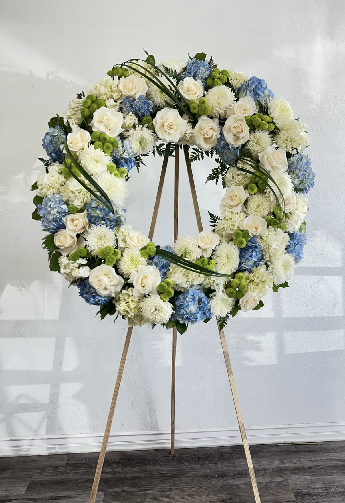 Blue Serenity Wreath  -  The Blue Serenity Wreath is a graceful tribute crafted with soft ivory roses, delicate white chrysanthemums, and serene blue hydrangeas. Accented with fresh green button poms and lush foliage, this elegant arrangement symbolizes peace, comfort, and eternal remembrance. Perfect for honoring a loved one, its soothing blend of blues and whites brings a sense of calm and heartfelt sympathy to any service or memorial.