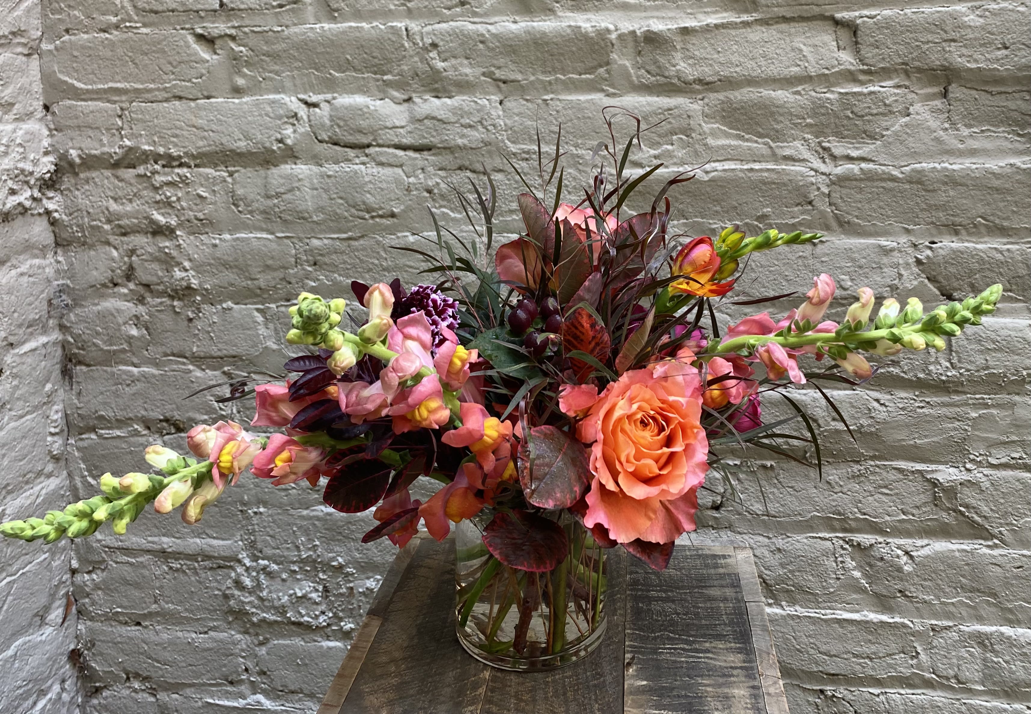 By the Bonfire  - A vibrant autumnal arrangement of vivid Free Spirit roses and fiery snapdragons set against luscious dark smokebush foliage.  Standard arrangement measures approximately 10x10&quot;.