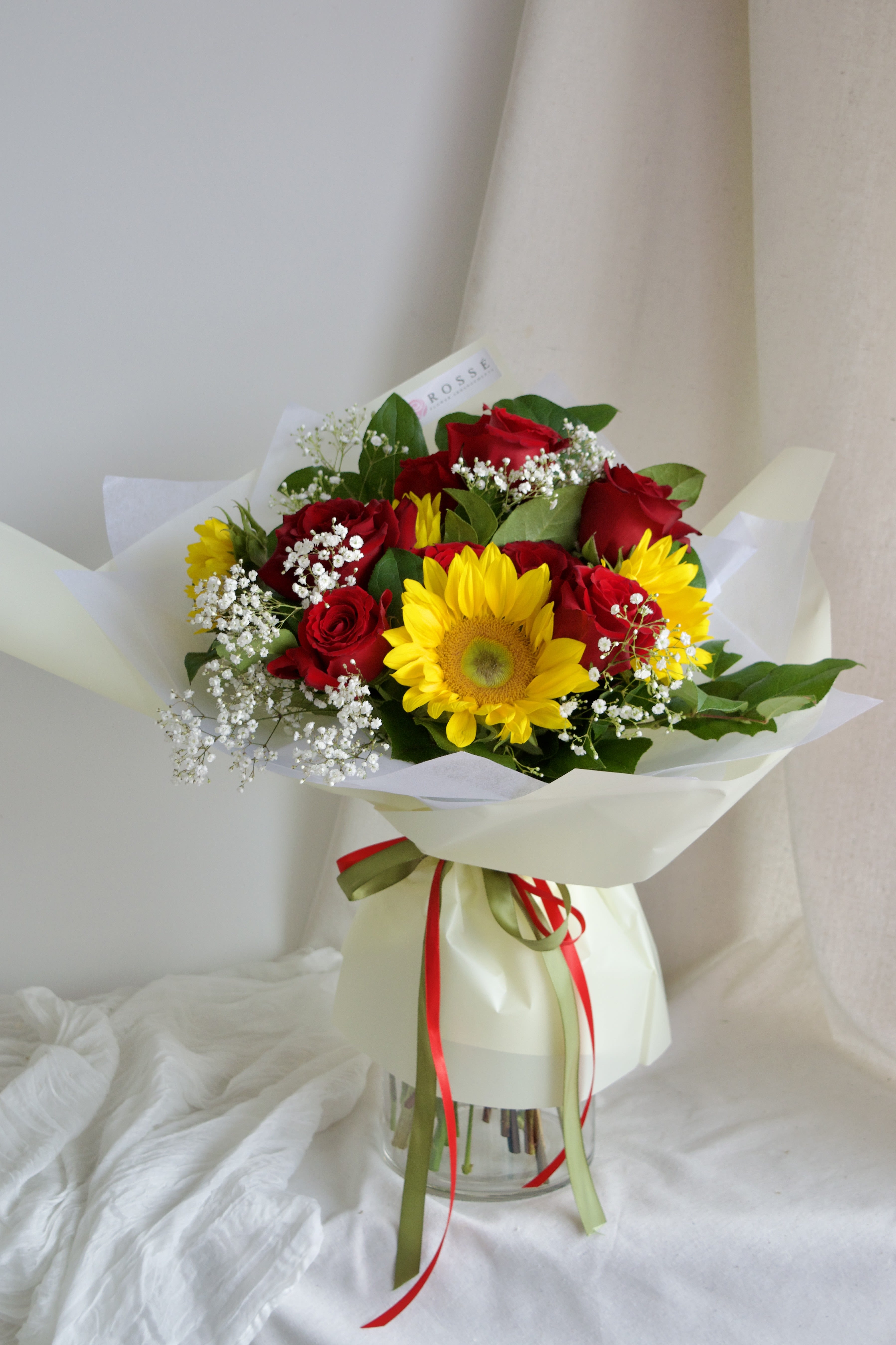 Sunset Love - A round bouquet with red and yellow flowers, greenery, wrapped in pastel yellow paper. Flower types may vary depending on the season.