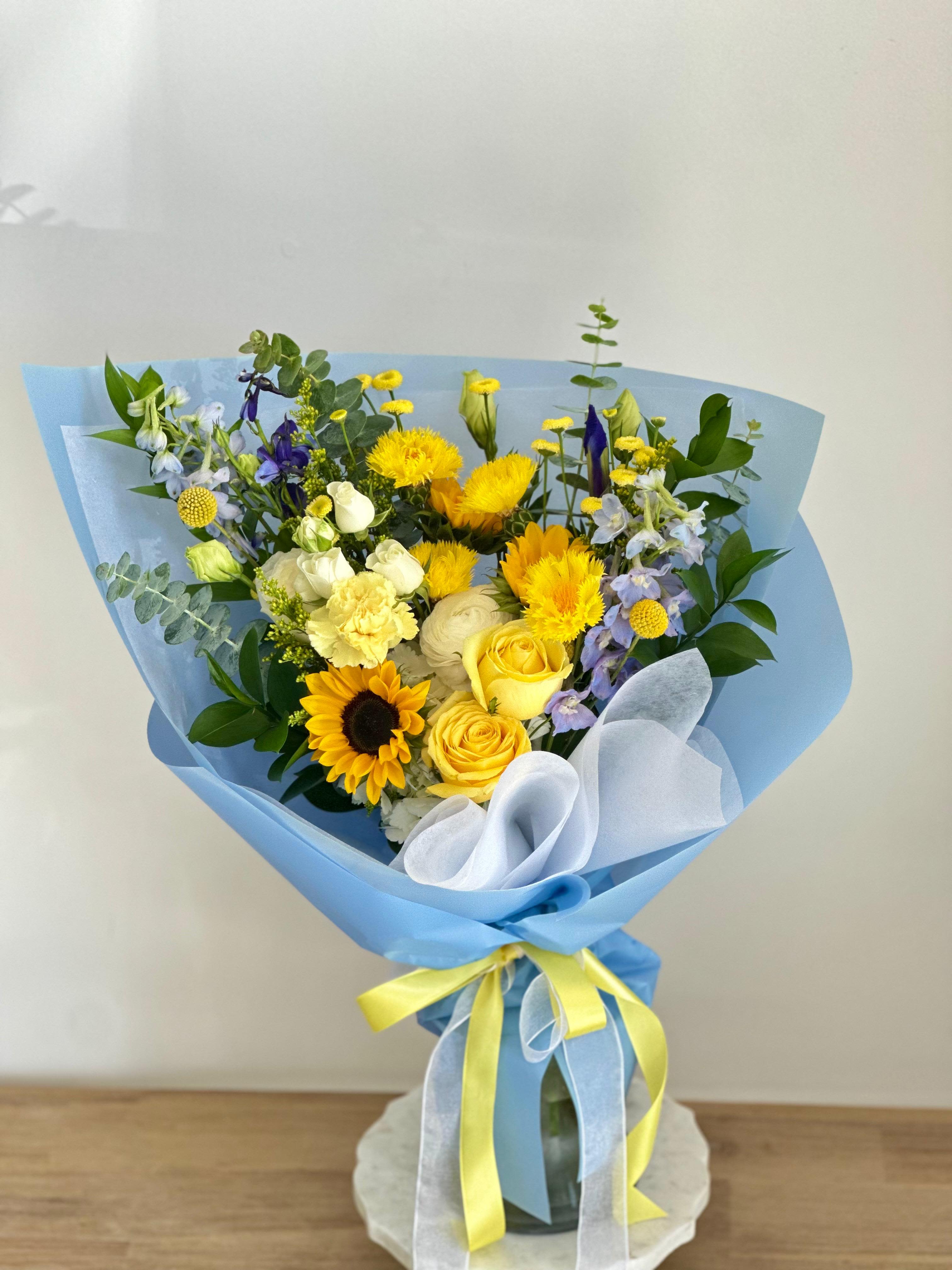 UCLA  - A front-facing bouquet with yellow, white, and blue flowers, wrapped in blue paper.   Flower types may vary depending on the season.  
