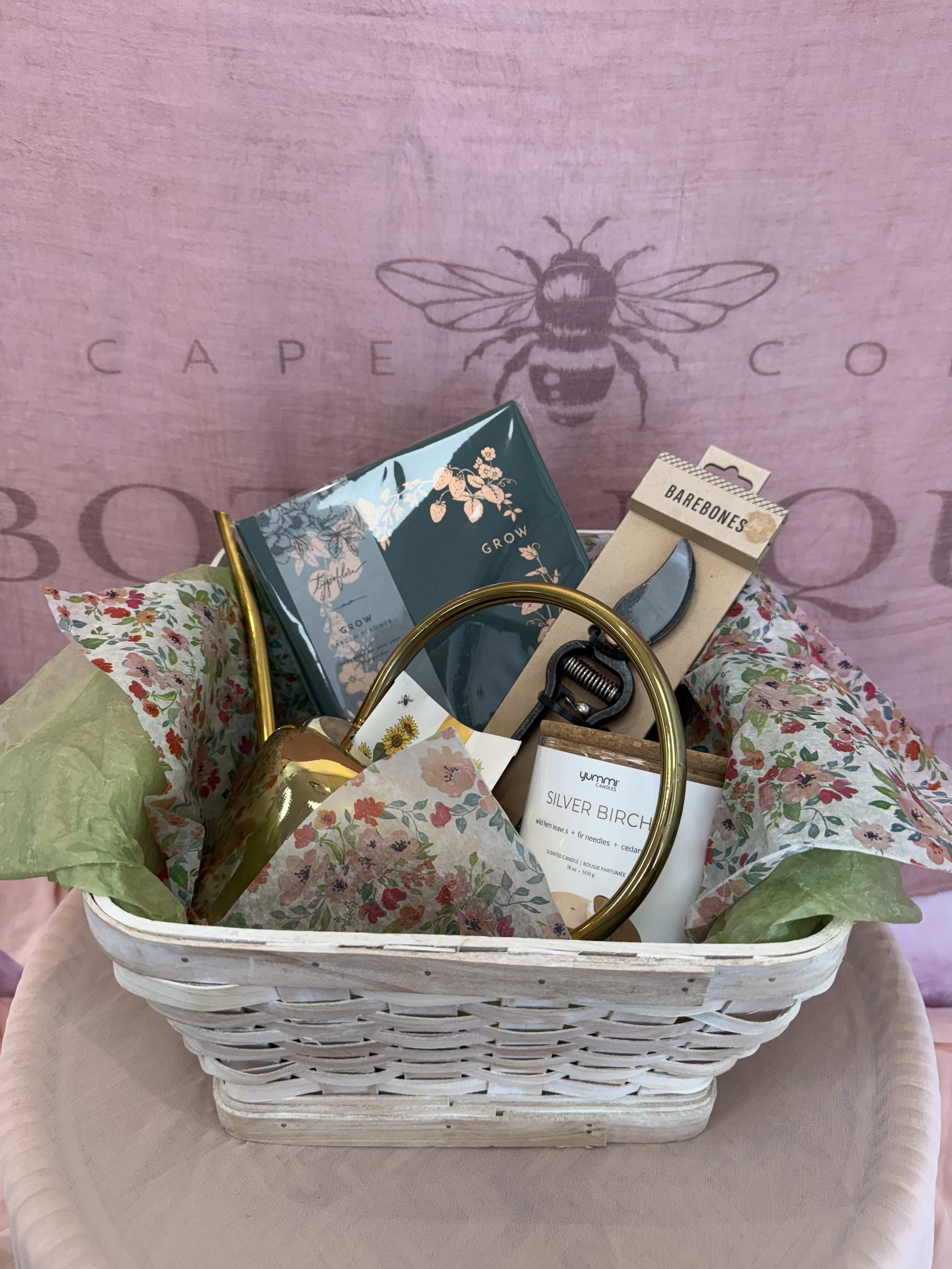 The Gardeners Companion Gift Basket  - A thoughtful collection designed for those who find joy in the garden. This charming basket includes a garden planner to map out blooms and harvests, sturdy pruners for perfect cuts, a watering can for nurturing growth and seed packets to start something new. To end the day, light the silver birch scented candle to unwind. Whether they're seasoned green thumb or a budding plant lover, this gift is the perfect companion for a season full of growing and blooming.   Gift basket includes:  1 Gold Henri watering can  1 Barebones, pruner   1 Typoflora - grow, garden planner 1 Yummi, Silver Birch Candle - 18oz. Plant based wax 1 Anellabees - sunflower seeds