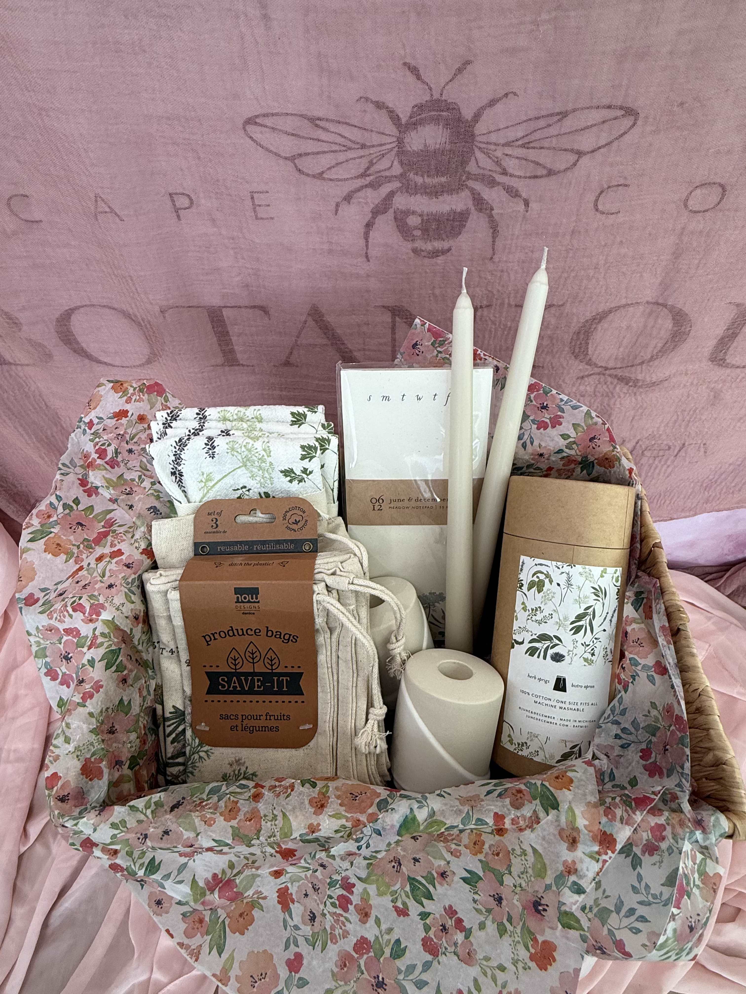 The Herb Garden Gift Basket - Infused with botanical charm, this gift brings the beauty of the garden to the table. It includes a set of four herb patterned cloth napkins, a coordinating bistro apron, a herb inspired notepad for jotting recipes or garden notes, and three reusable produce bags adorned with herbs. To complete the ambiance, two taper candles with elegant ceramic holders add a soft, cozy glow - perfect for slow dinners and fresh beginnings in the kitchen.   Gift Basket includes:  4 June &amp; December Herb Springs napkins 1 June &amp; December Herb Springs Bistro Apron 1 June &amp; December Herb Notepad  1 Now designs by Danica, Save It - Herb produce bags 3 pack  ( 3 sizes: 8 x 10 / 10 x 13 / 12 x 16) 2 Lenne candle holder  2 Camilia Candle 12&quot;, Ivory