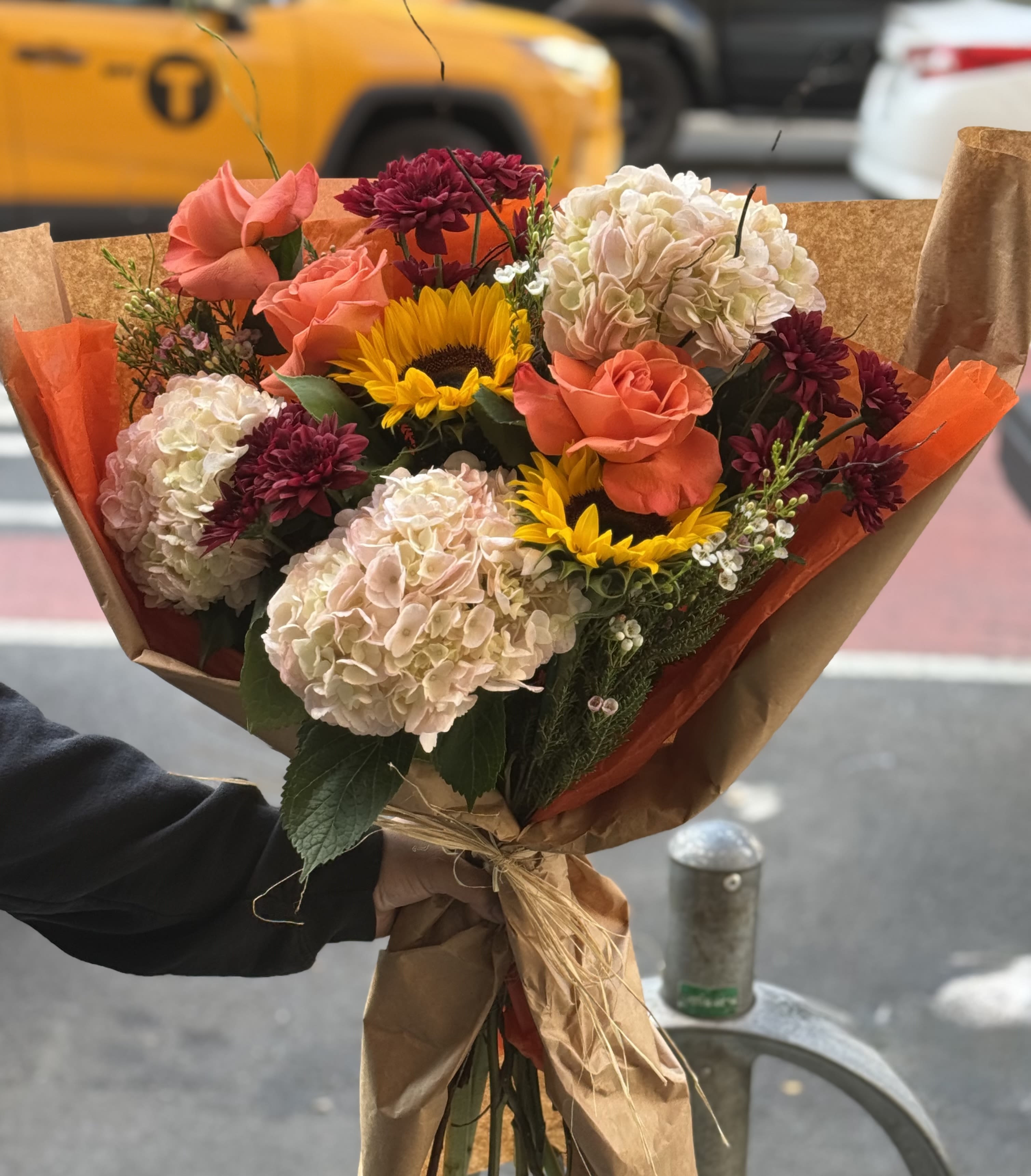 Autumn Glow - A warm and cheerful hand tied bouquet featuring vibrant oranges and sunny yellows. Accented with delicate fillers and wrapped in rustic brown and orange paper - perfect for any fall celebration or just to brighten someone’s day.   Note: No vase included.