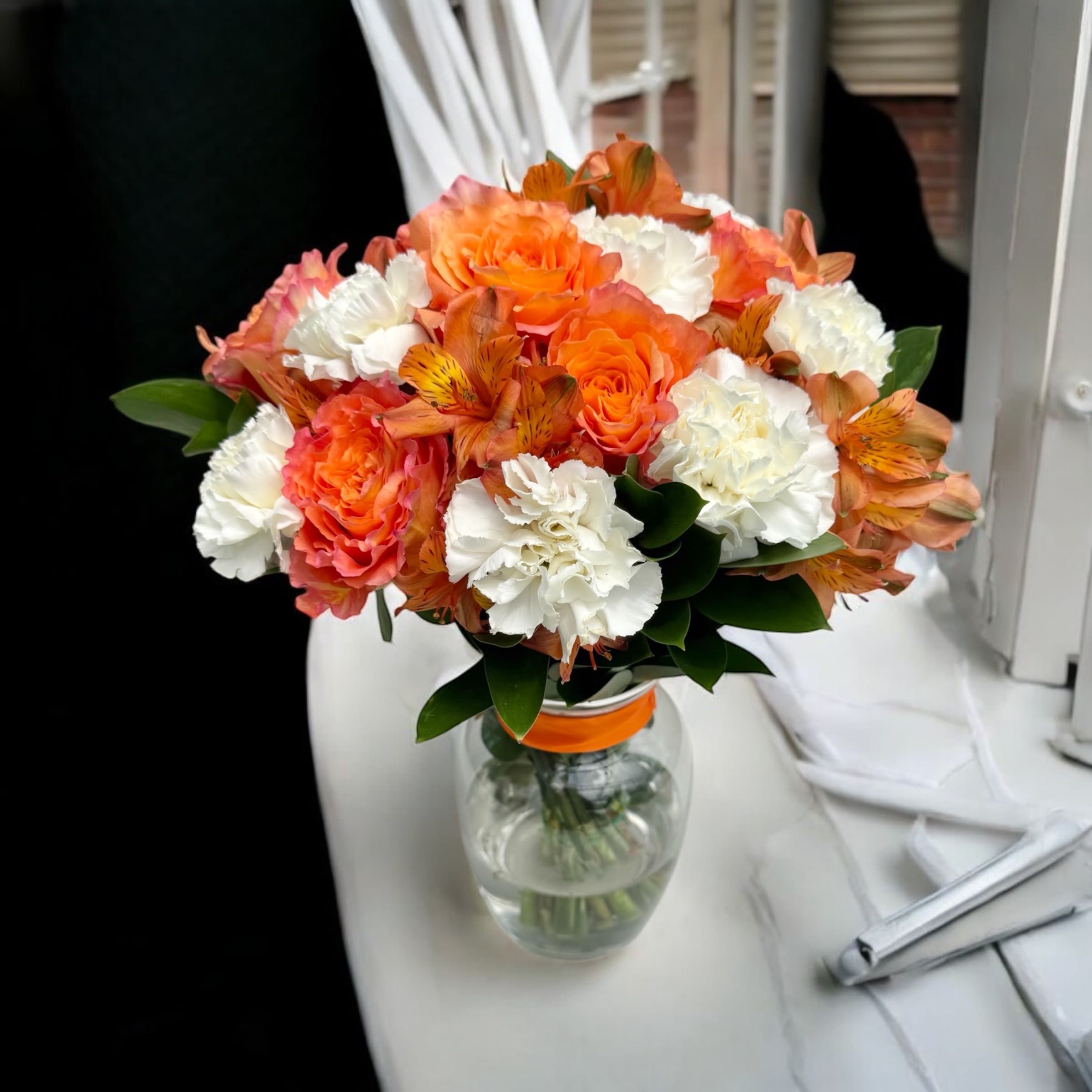 Sweet Emotion Citrus MDay - Radiate warmth and charm with our Orange Rose and White Carnation Bouquet. The vibrant orange roses, paired with the elegance of white carnations and the delicate touch of baby's breath and Alstroemeria, create a striking and inviting arrangement. Surrounded by lush greens, this bouquet captures the essence of joy and happiness, making it a perfect gift for any occasion.