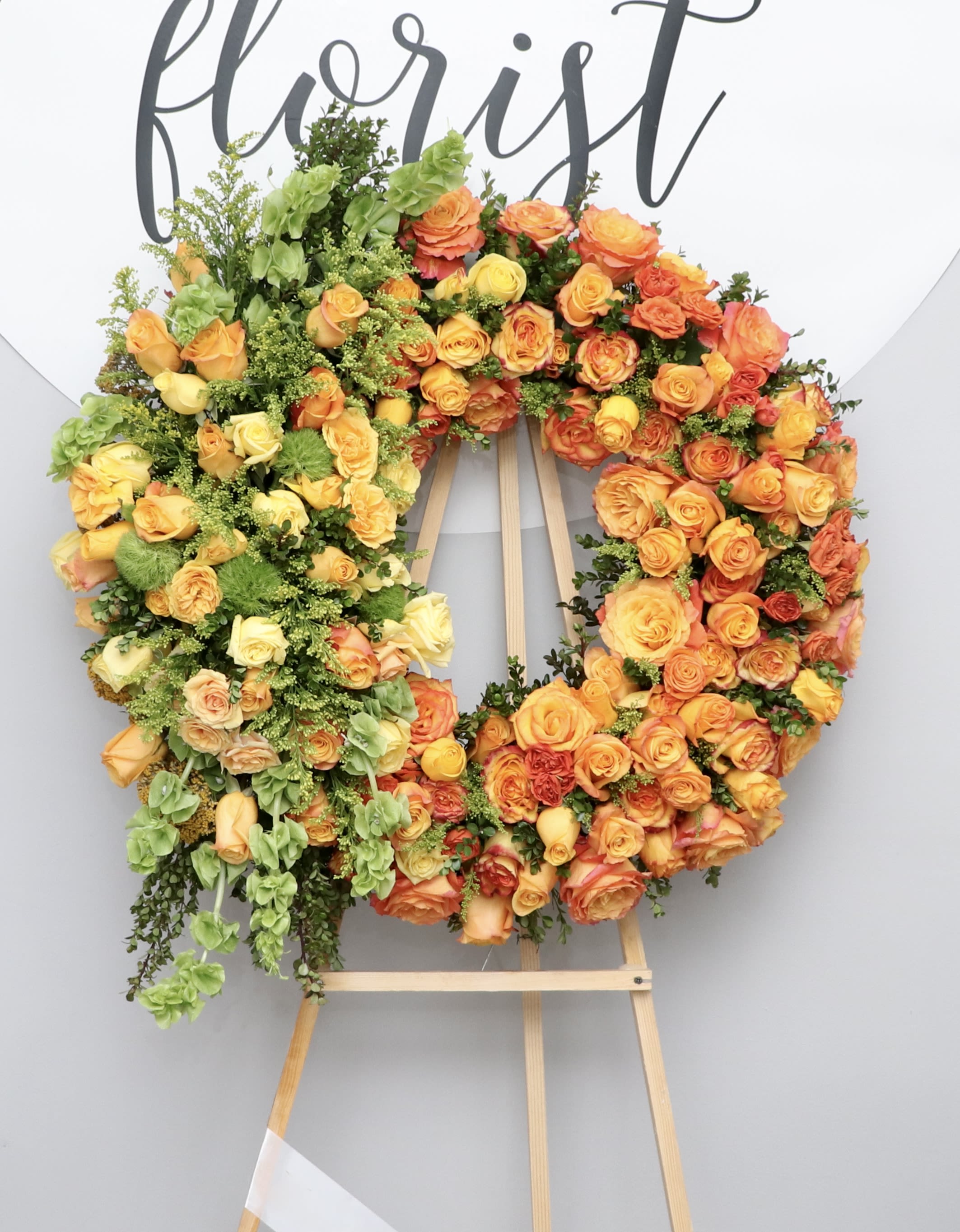Sunlit Farewell  - The Sunlit Farewell is a warm and elegant funeral wreath, featuring orange and yellow roses and seasonal leaves. The combination of colors and textures create an inviting and comforting atmosphere that celebrates the life of the departed.  We include easel, printed banner, and delivery (some fees may apply). An additional banner can be added for $15  Standard size is 30'', deluxe is 36'', and premium is 42''.
