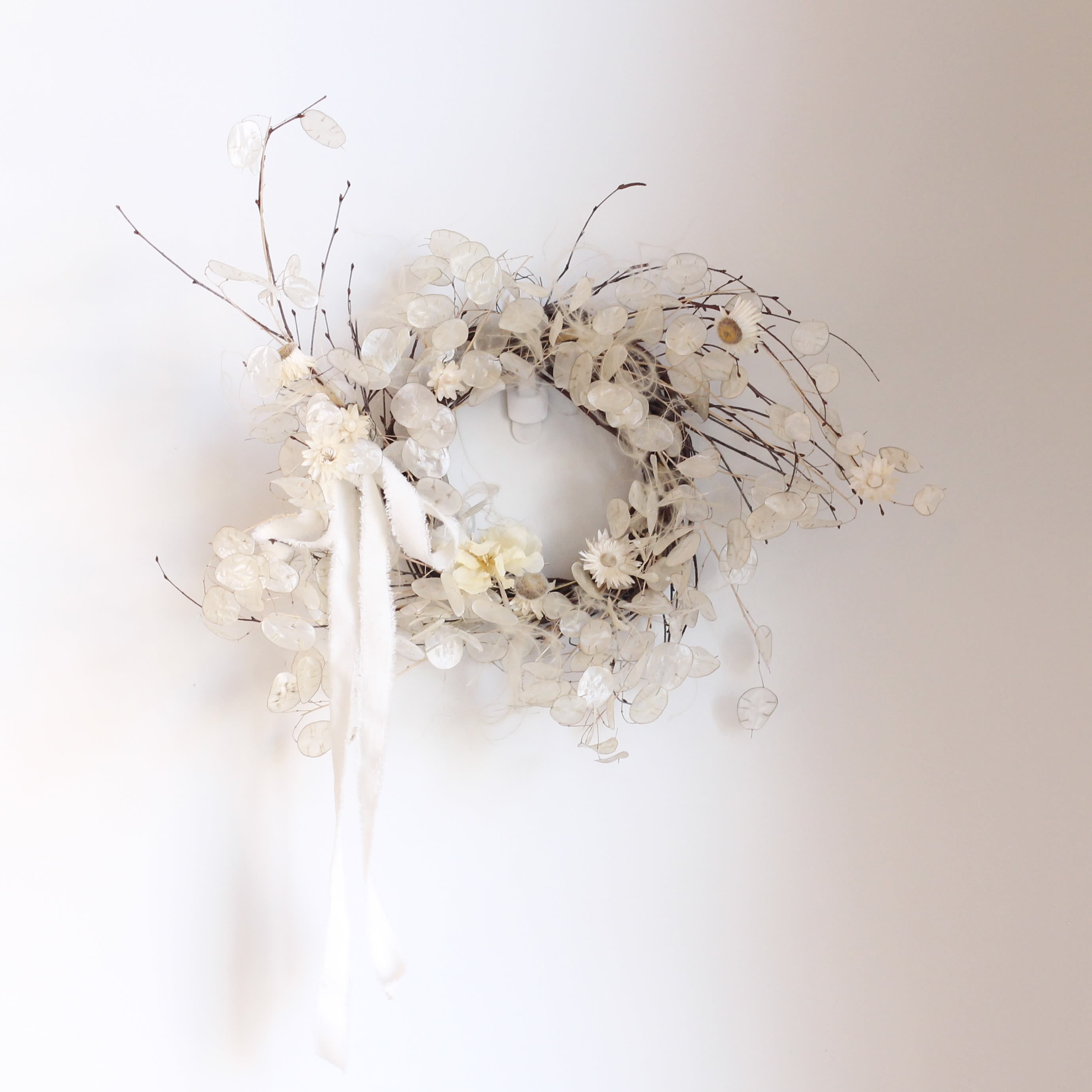 &quot;Lunaria Nest&quot; Dried Floral Wreath - Delicate and ethereal, this handcrafted wreath features shimmering Lunaria also known as “honesty” or “silver dollar” plant whose translucent seed pods catch the light with a soft, moonlike glow. Wisps of troll hair add whimsical texture and movement, while clusters of preserved strawflowers bring subtle color and charm. Finished with a white velvet ribbon, this wreath blends natural beauty and elegant simplicity, perfect for brightening any space year round. Appx 20&quot; across 