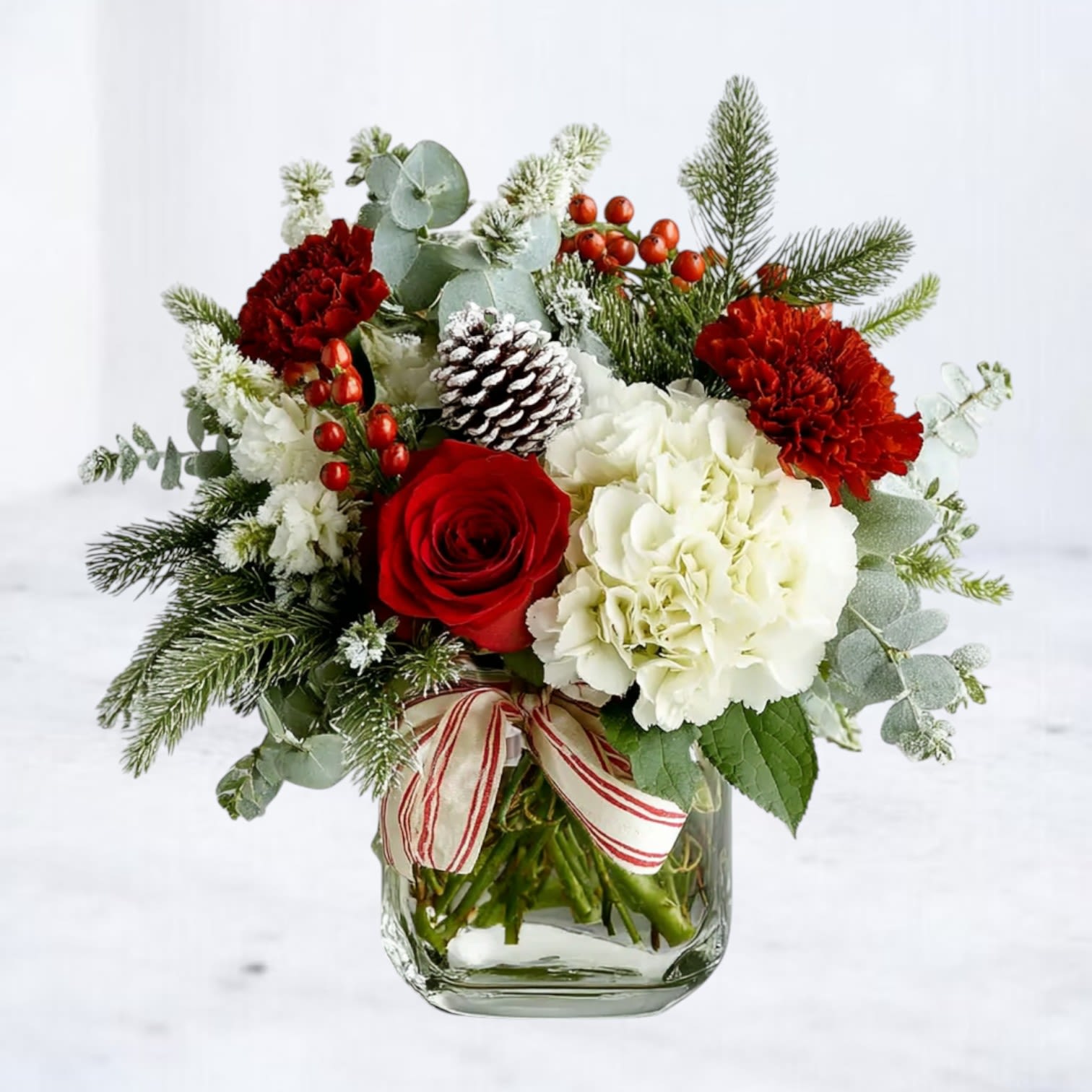 Scarlet Hearth - The Scarlet Hearth arrangement captures the timeless charm of the holiday season with rich red roses, snowy white hydrangea, crimson carnations, and frosted evergreens. Accented with pinecones, berries, and a festive striped ribbon, this design brings the warmth of the holidays into any home.  Crafted in a clear glass vase, it’s both classic and welcoming — a perfect centerpiece or gift that feels like home for the holidays.