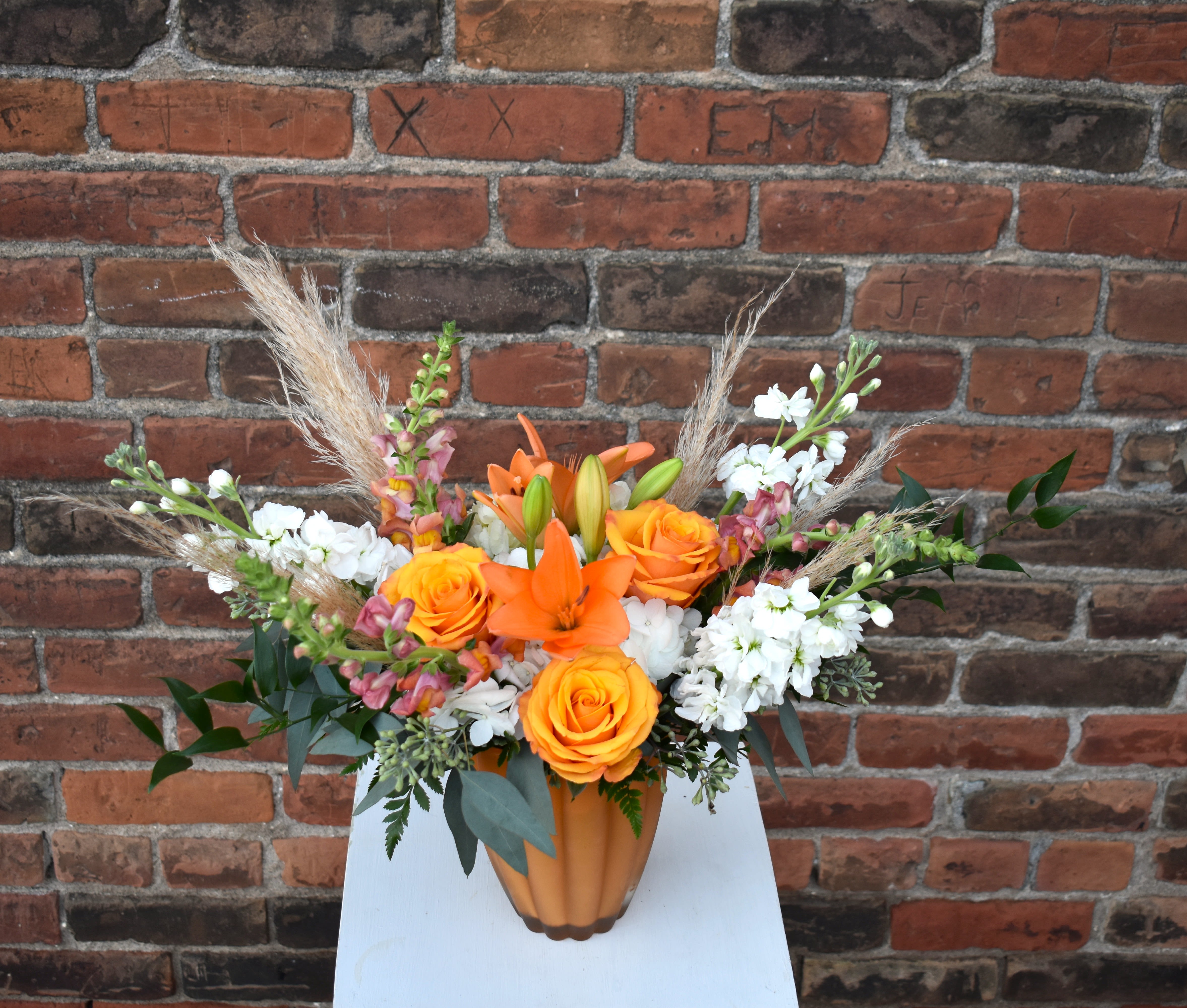 Pumpkin Pie - A delightful fall bouquet brimming with vibrant orange snapdragons, roses, lilies, white hydrangea, stock and lush greenery designed in an orange scalloped vase and accented with rustic pampas grass. A perfect treat for celebrating the season! 