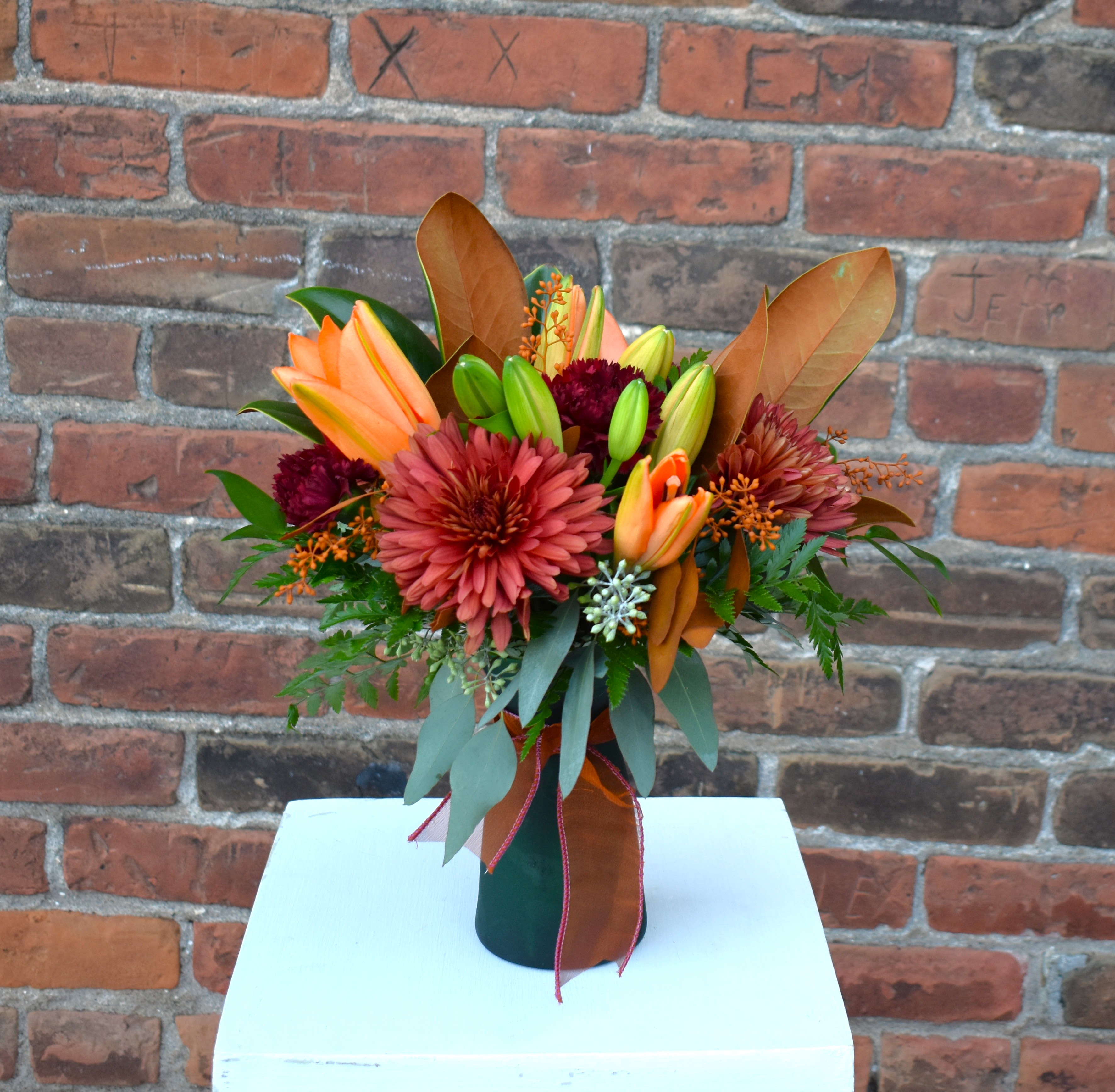 Amber and Emerald - A warm, rustic bouquet designed with terracotta mums, burgundy carnations, bright orange lilies, mix of eucalyptus, accents of magnolia in a emerald vase detailed with copper ribbon...a toasty way to celebrate the season! 