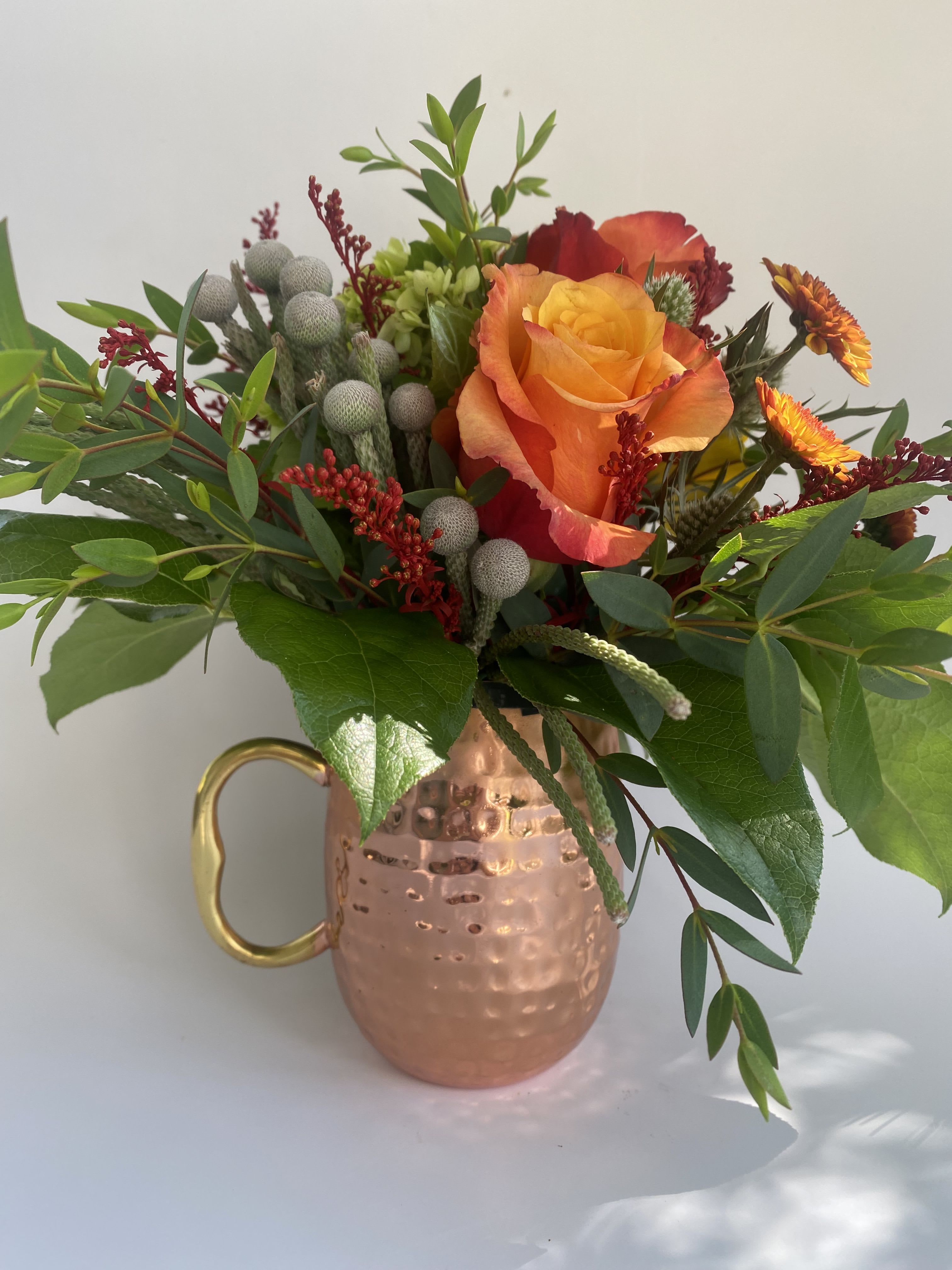 Autumn Cup of Cheer - Celebrate the Warmth of Fall with a Unique Twist Introducing &quot;Autumn Cheer,&quot; a distinctive arrangement that captures the essence of fall in a charming copper mug, reminiscent of cozy apple cider moments or the classic &quot;Moscow Mule&quot; cocktail. This arrangement is overflowing with fresh cut, fall-hued florals including roses, mini-green hydrangeas, seasonal fillers, mums, thistle, and berries. Fall-Hued Splendor: A vibrant mix of autumnal colors brings warmth and joy, perfect for celebrating the beauty of the season. Unique Copper Mug: The copper mug adds a rustic and festive touch, doubling as a keepsake for future cozy moments. Versatile Gift: With its rich, earthy tones and unique presentation, this arrangement is a fitting choice for anyone who loves fall, including the fellas. &quot;Autumn Cheer&quot; from Elizabeth's Garden is the perfect way to share the joy and richness of the fall season, offering a unique and memorable gift for any occasion.