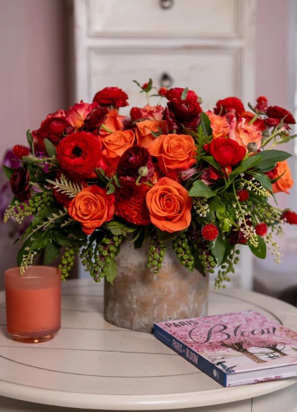 Autumn Sunset - This fiery floral arrangement is a vibrant autumn hue. Rich, velvety red and burgundy ranunculus intertwine with sun-kissed Orange Crush and Free Spirit roses, creating a captivating contrast of color. The exotic protea amplifies this piece’s beauty and drama. Finally, the delicate spikes of coxcomb provide a playful texture and complete this masterpiece.  Orientations: One-sided.